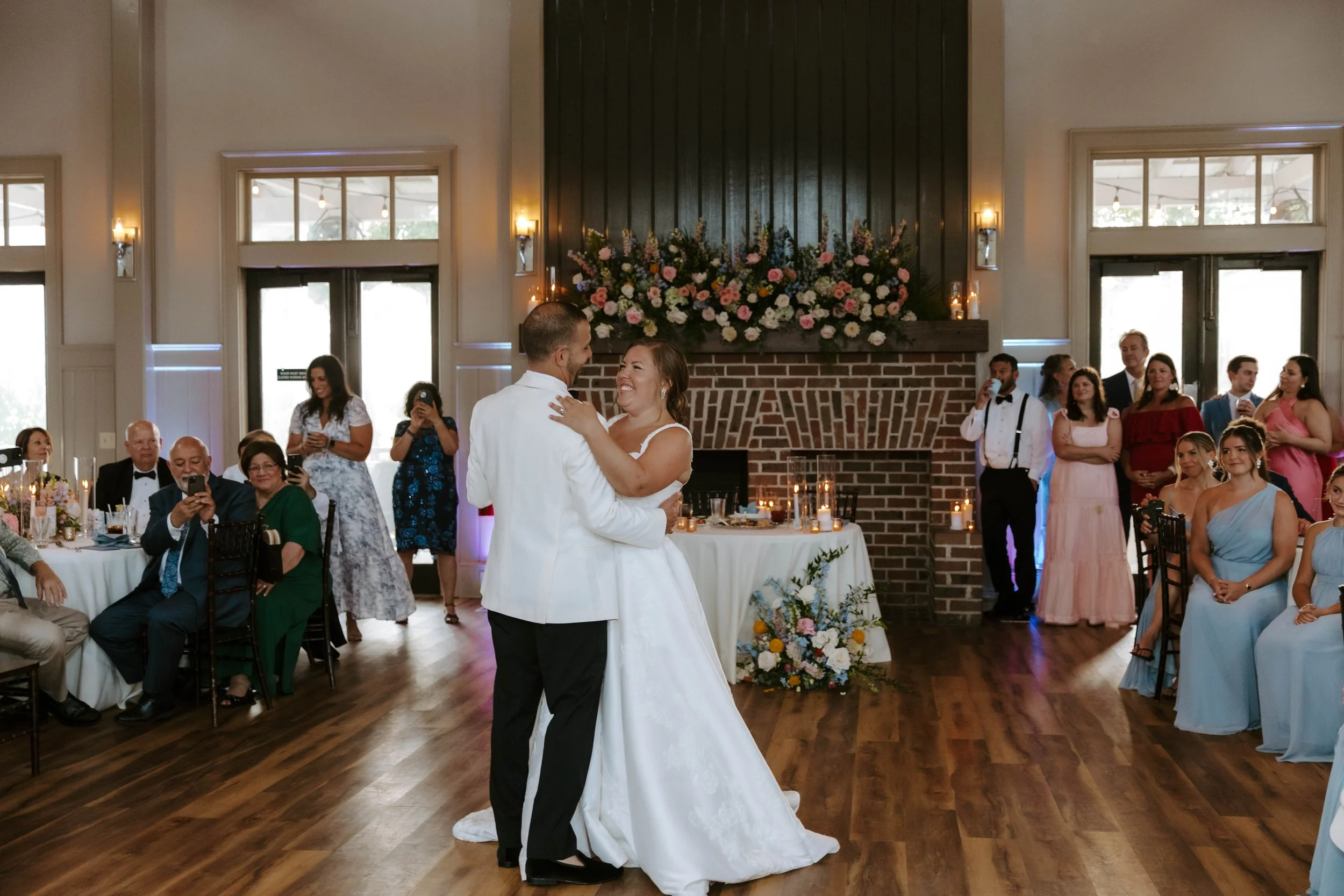 A bride and groom are dancing together at their wedding reception. Guests are seated and standing around, taking photos and watching. The room has high windows, a brick fireplace with flowers above, and warm lighting.