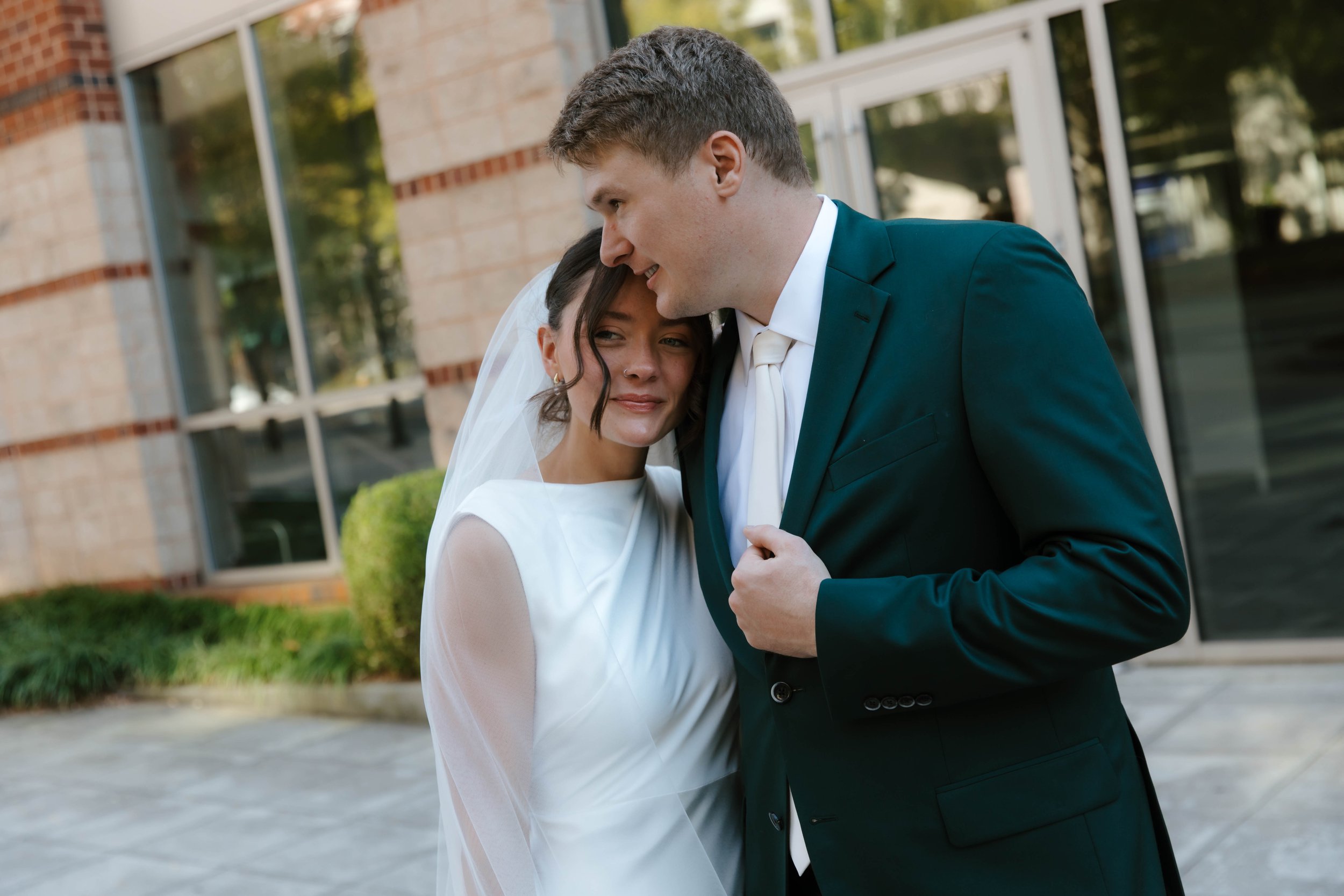 A bride and groom sharing an intimate moment outside a building with large glass windows. The bride is leaning her head on the groom's chest and smiling softly, while the groom is looking down at her with a gentle expression. They are dressed in wedding attire, with the groom in a dark suit and the bride in a white dress and veil.