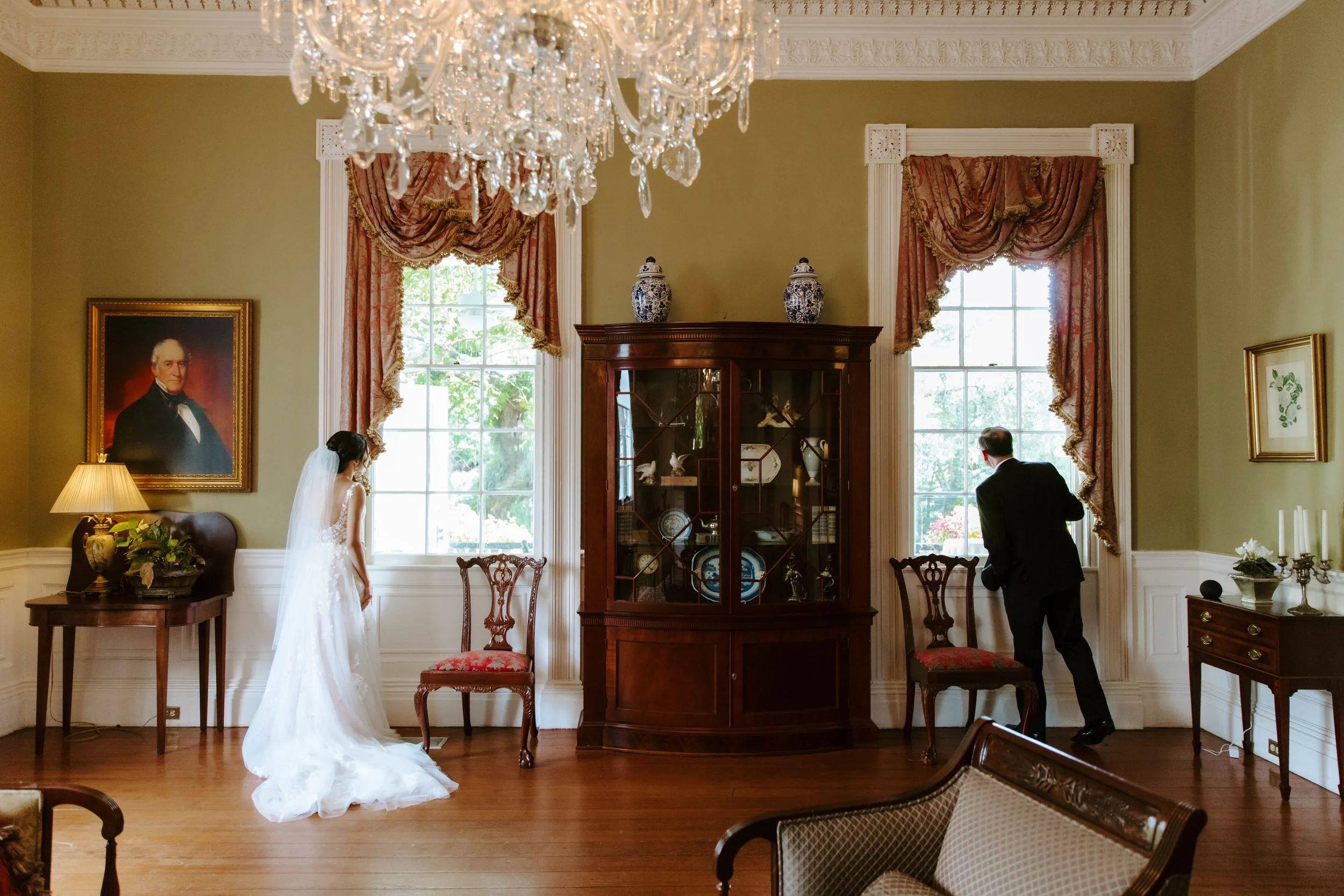 A bride and groom in a decorated room with large windows, ornate curtains, a chandelier, a portrait painting, and antique furniture.