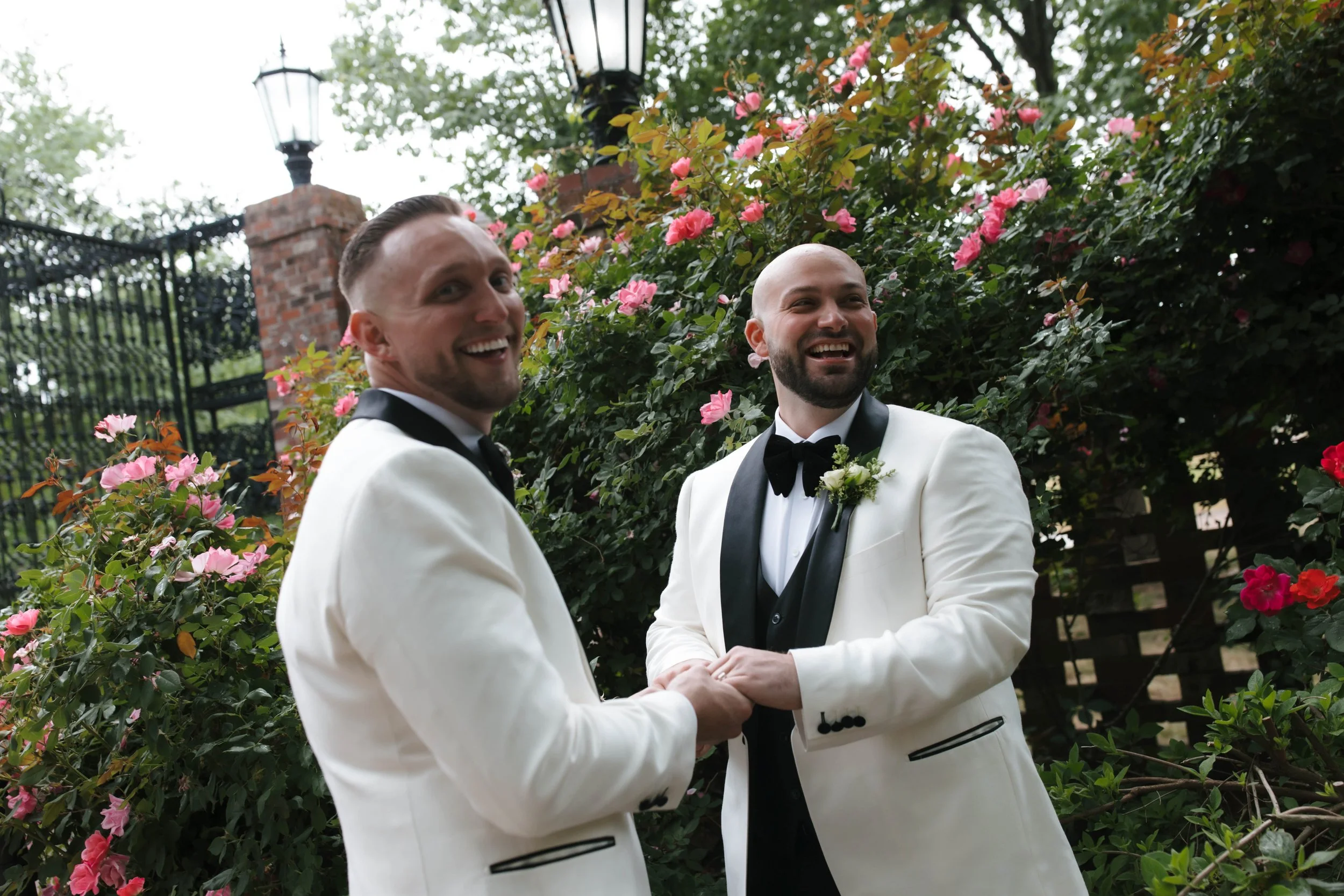 Two men in tuxedos smiling and holding hands in front of pink and red roses, with a black gate and street lamps in the background.