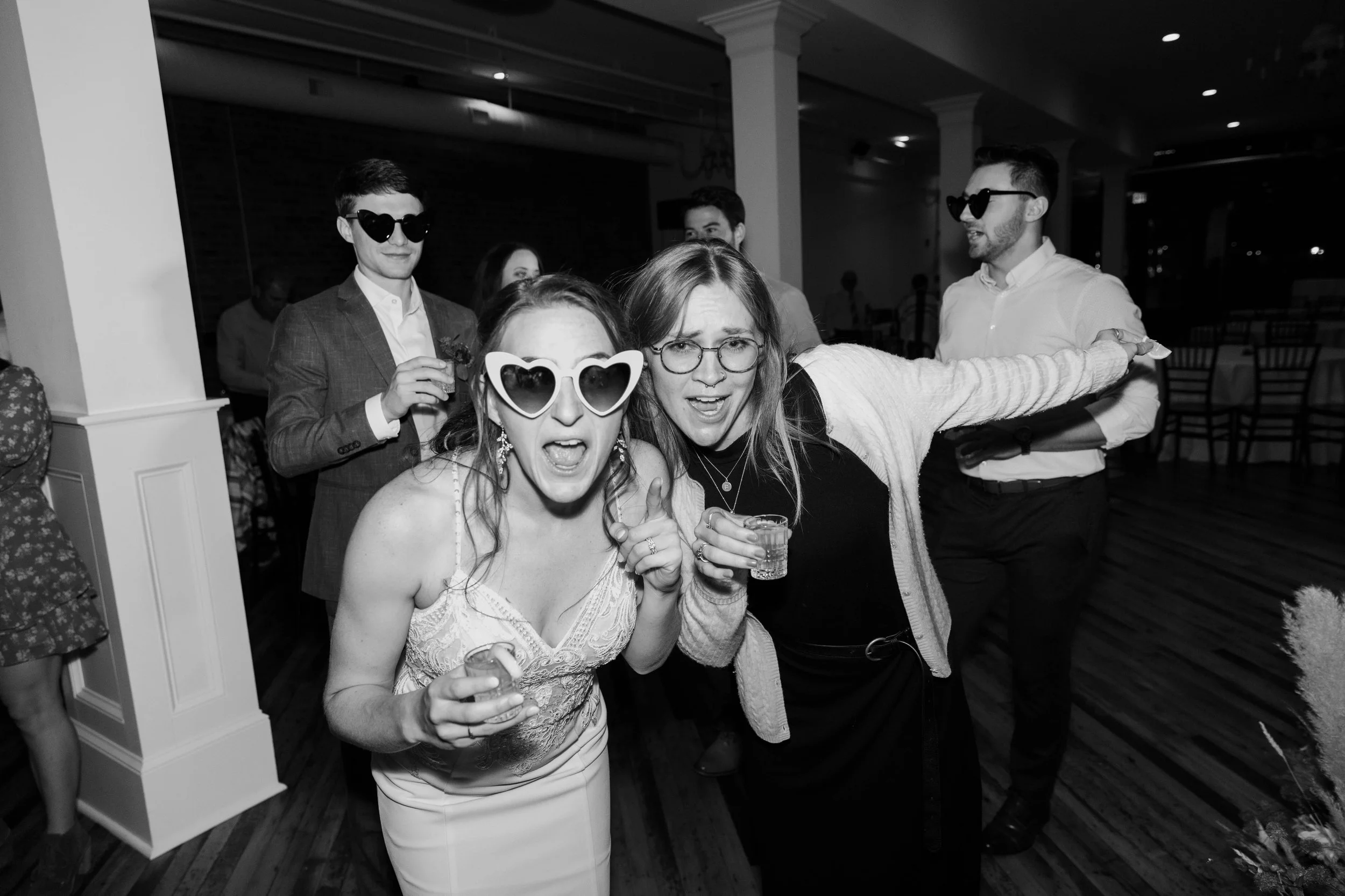 Group of people at a party, dancing and celebrating with one woman in heart-shaped sunglasses and others holding drinks, in a dimly lit indoor venue.