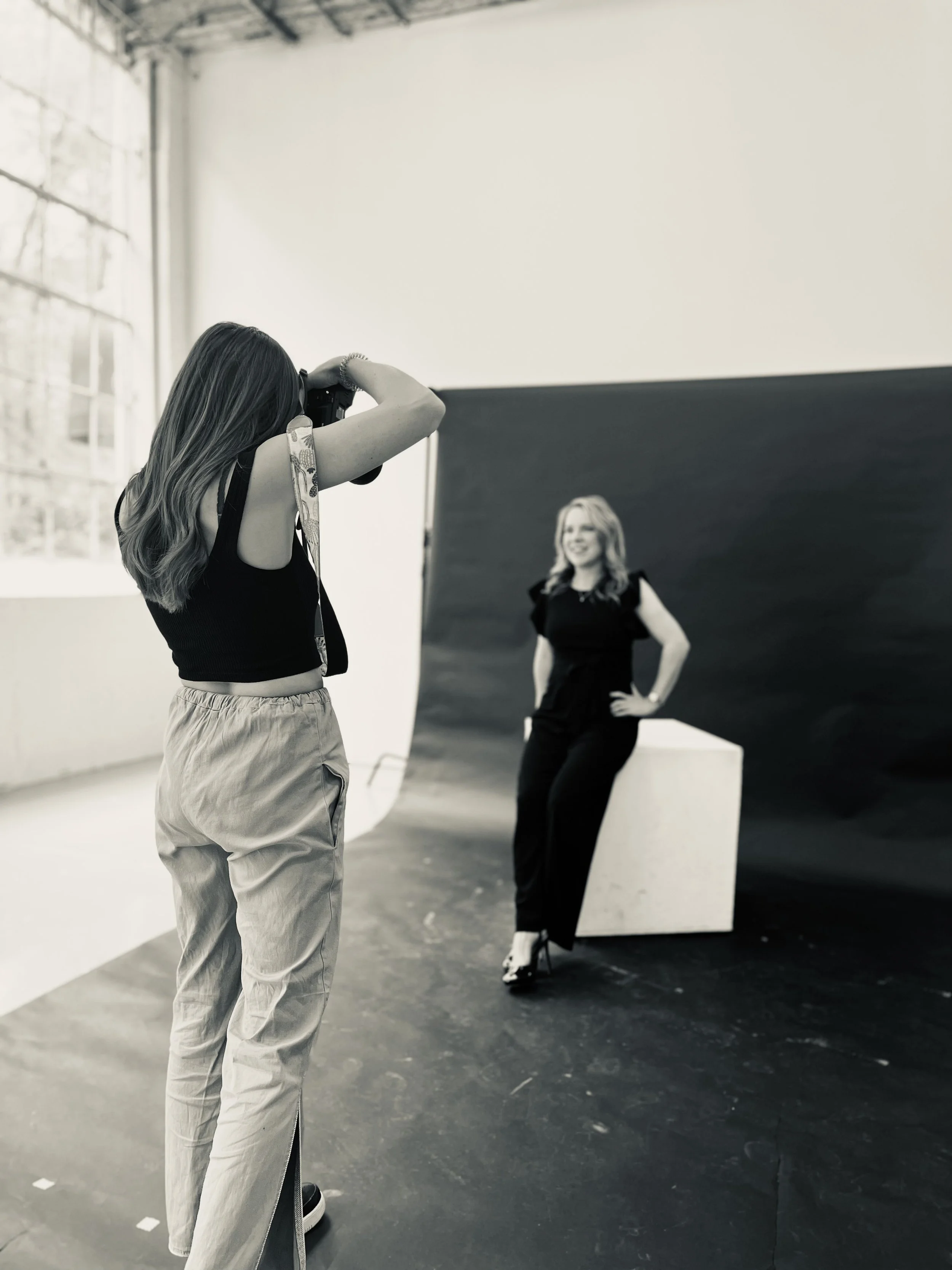 A woman with long hair wearing a sleeveless top and loose pants taking a photograph of a model posing in front of a black background in a photography studio.