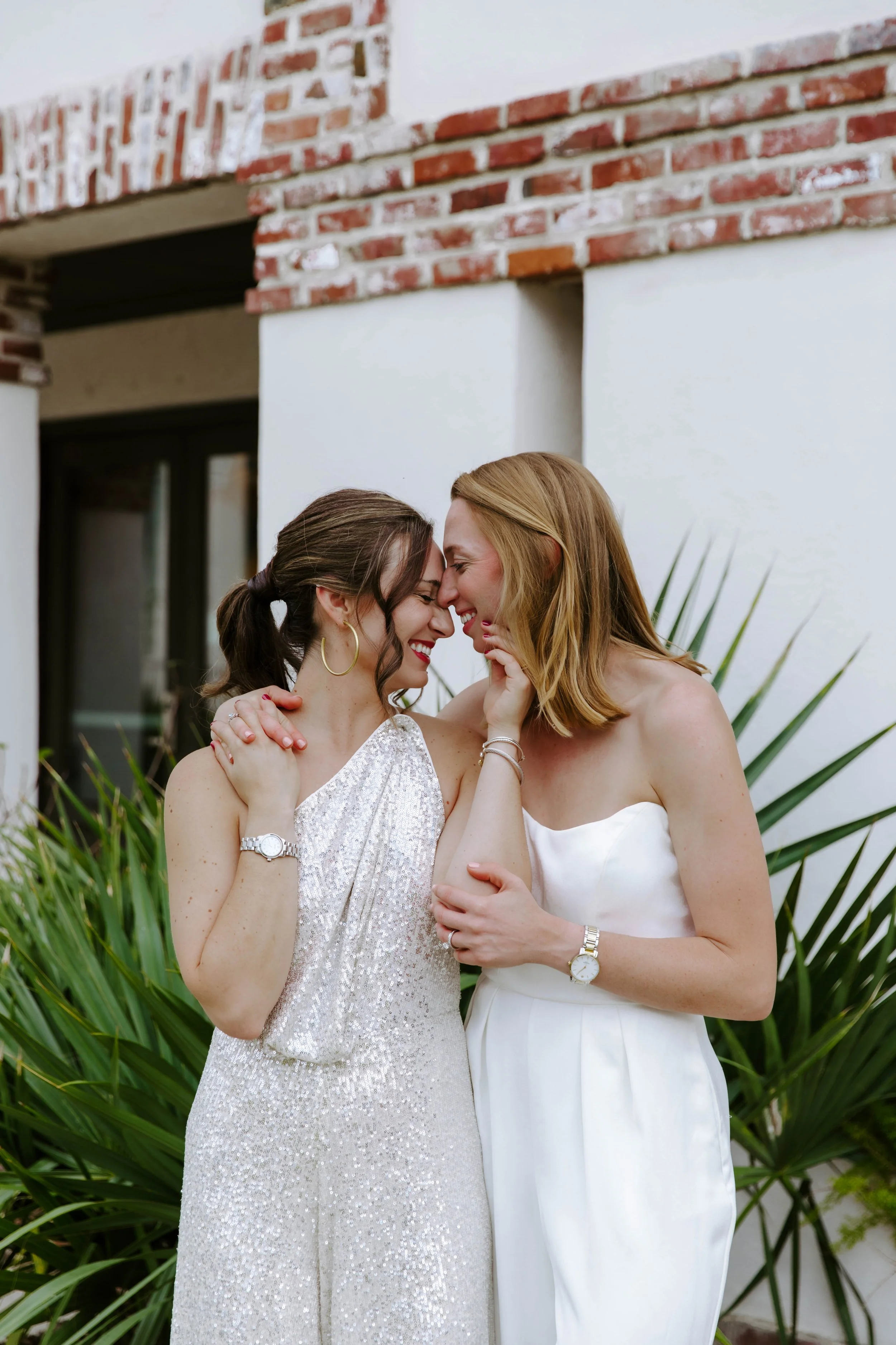 Two women are close together, touching foreheads and smiling. One is wearing a shiny, silver dress and the other a white dress. They are outdoors with green plants and a brick wall in the background.