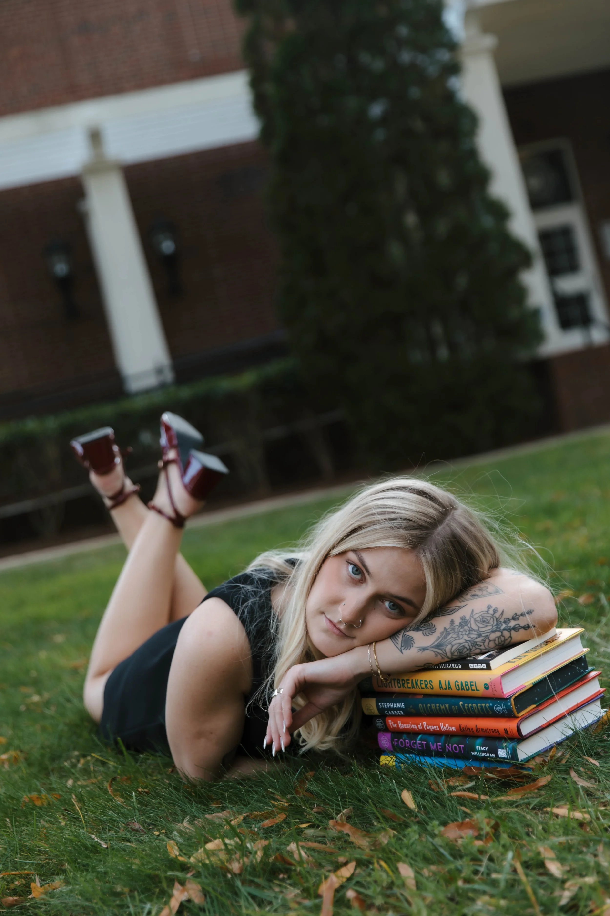 A young woman with blonde hair, tattoos, and a nose ring lies on her stomach on grass, resting her head on her arms, with a stack of books beside her. She is outdoors near a building with a brick wall, bush, and trees, and is wearing a black dress and burgundy sandals.