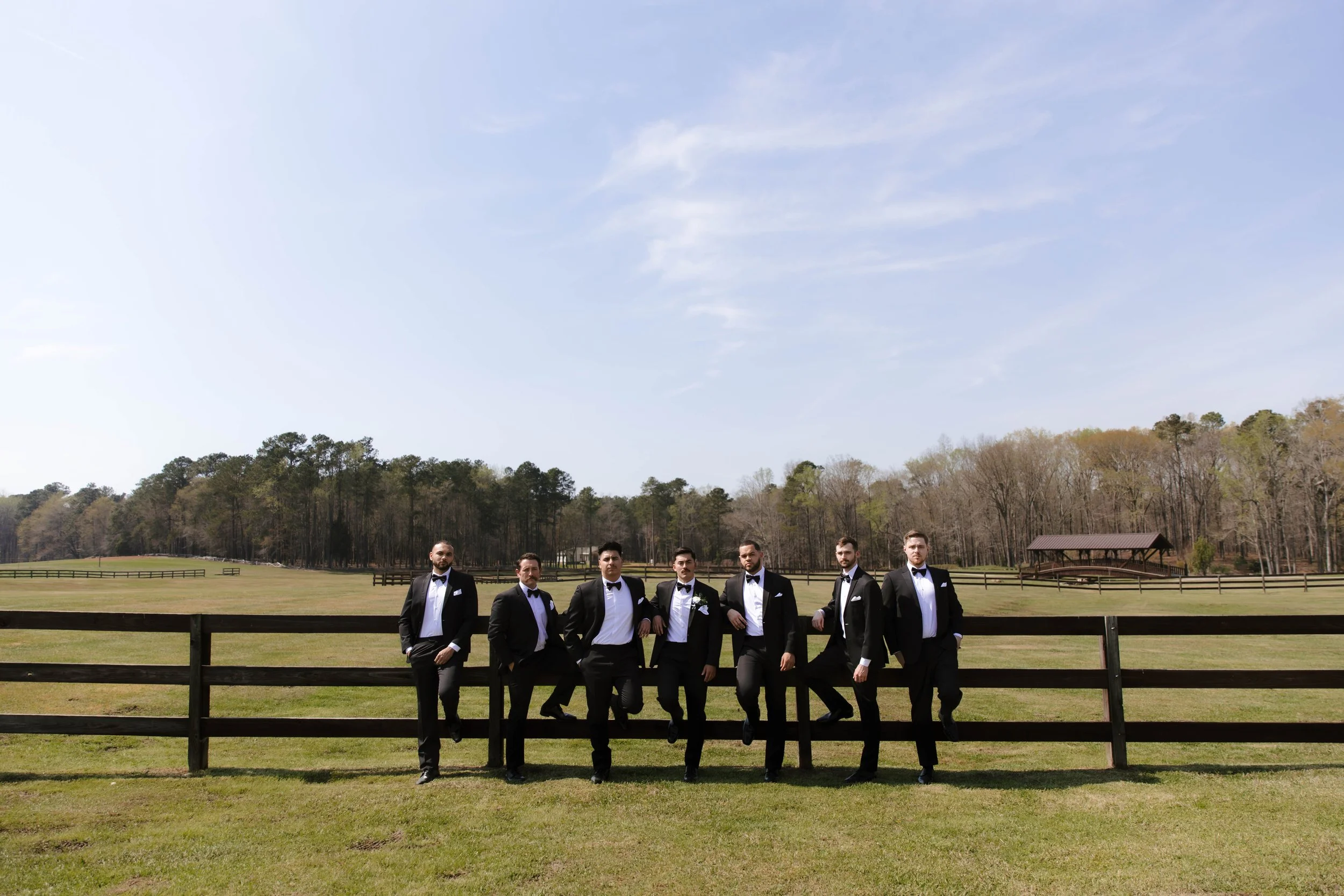 A group of eight men in tuxedos standing outdoors in front of a wooden fence, with a grassy field, trees, and a pavilion in the background, under a partly cloudy sky.