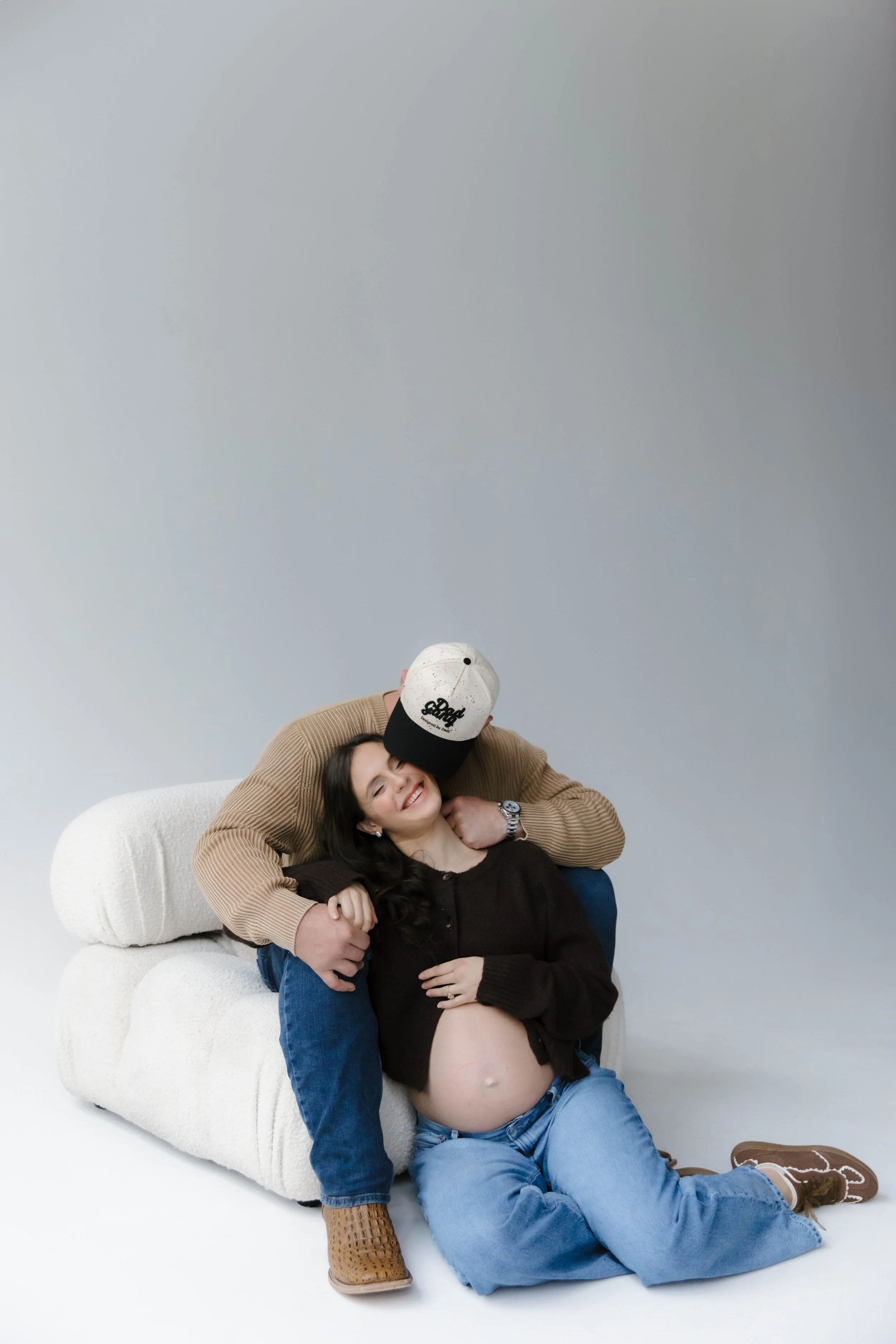 A pregnant woman sitting on the floor with a man behind her hugging her and a woman beside her smiling. The man is wearing a beige sweater, jeans, and a baseball cap, while the pregnant woman is wearing a black sweater and jeans, and the woman beside her is in a black outfit.