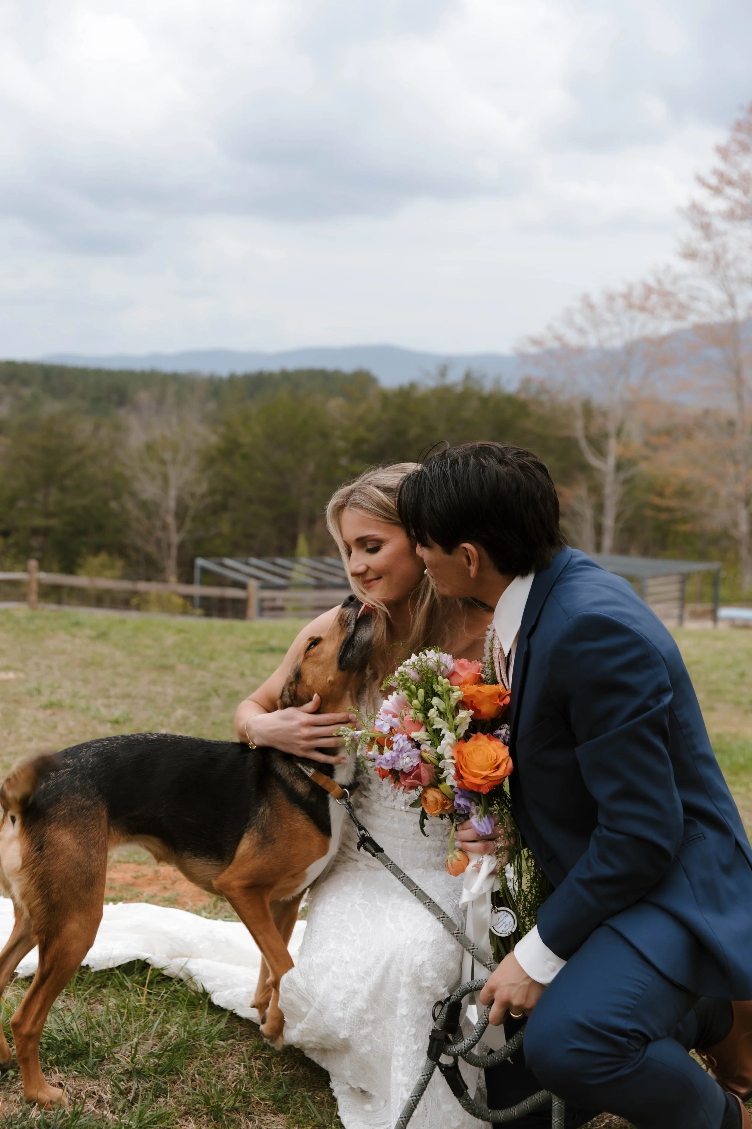 A bride and groom kneeling on grass, embracing and petting a dog during their outdoor wedding ceremony with mountains and cloudy sky in the background.