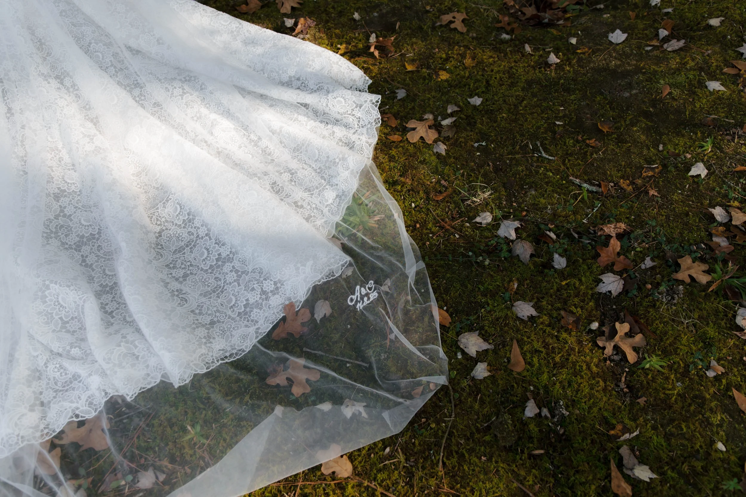 A white lace wedding dress with embroidered detailed floral patterns lies on mossy ground with fallen leaves.