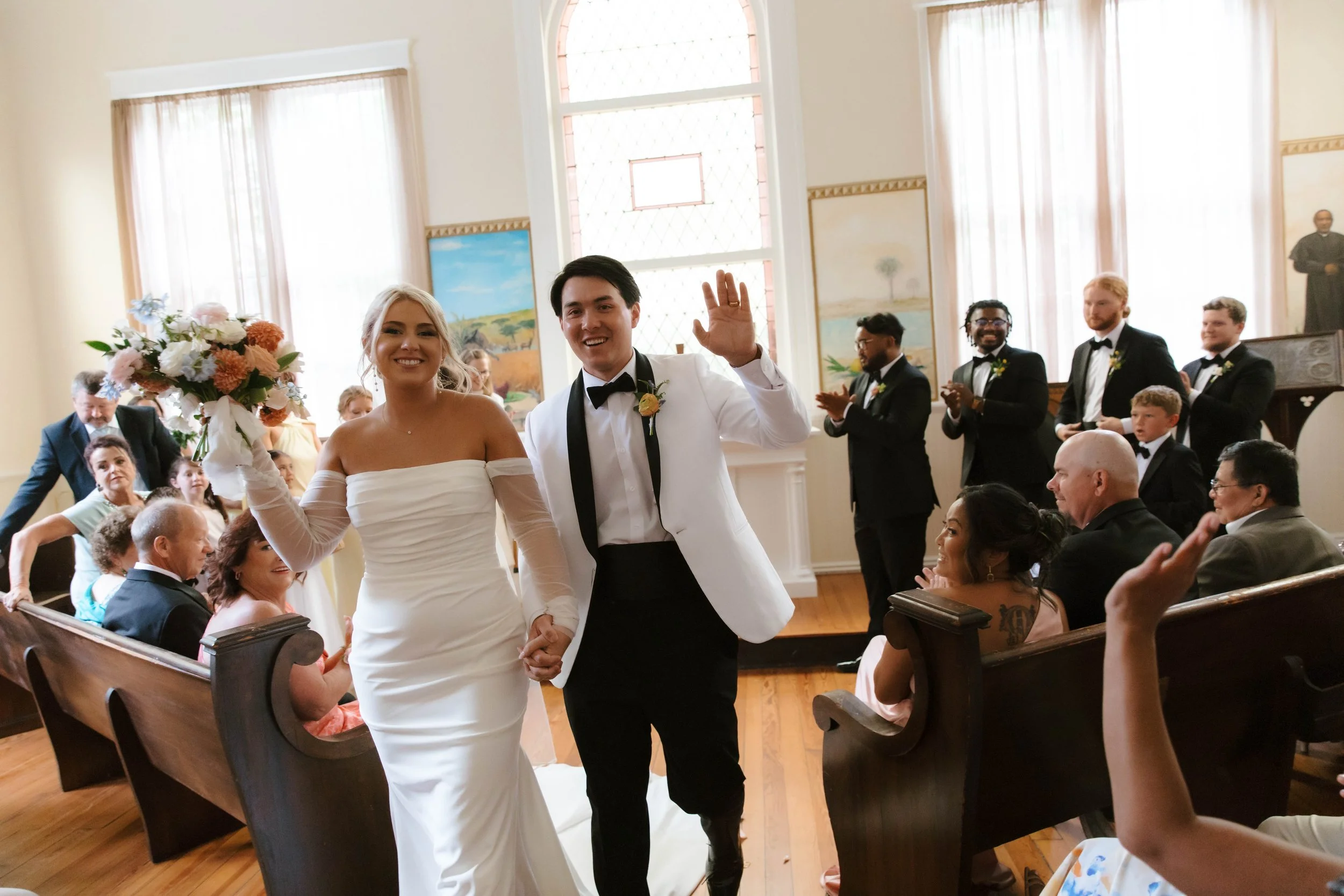 A bride and groom holding hands and smiling at their wedding ceremony in a church, surrounded by seated guests and standing groomsmen.