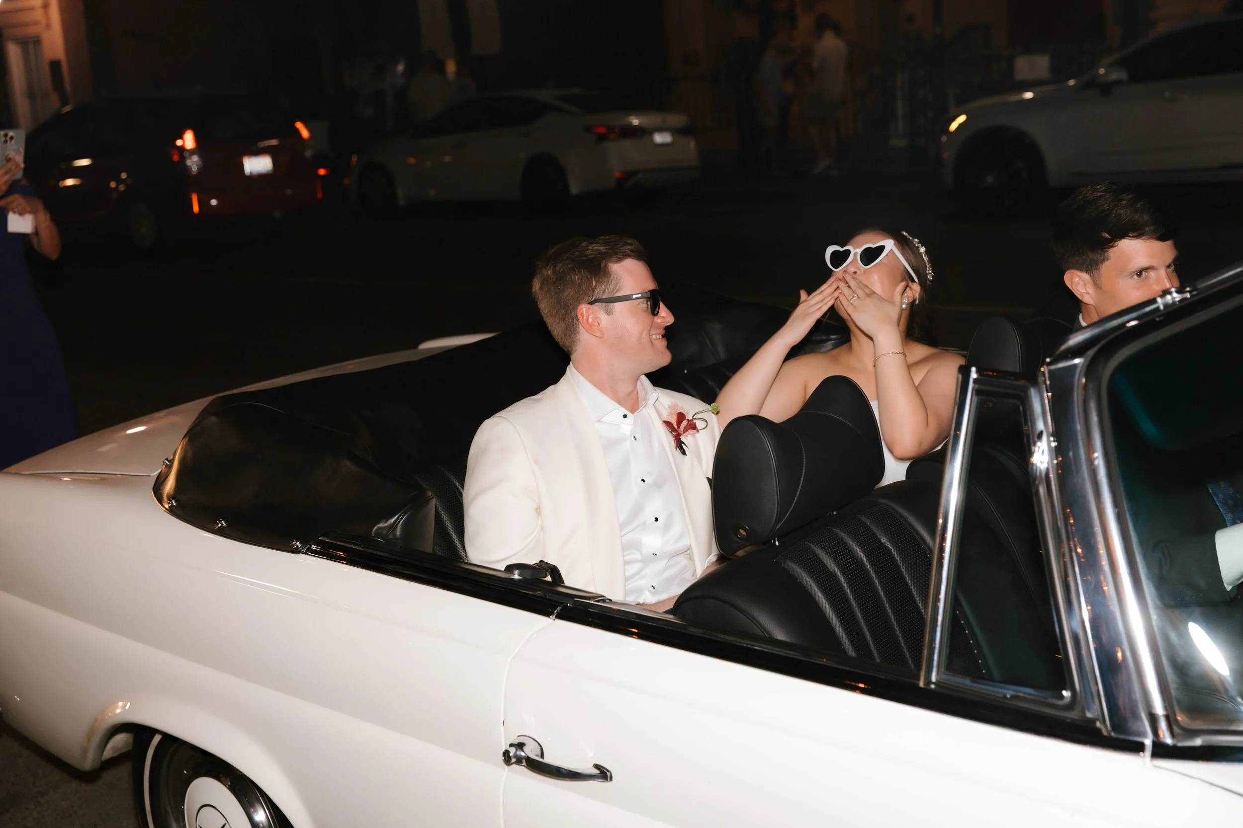 A bride and groom dressed in formal attire sitting in a white convertible car at night, sharing a joyful moment with the woman blowing kisses, wearing heart-shaped sunglasses, and the man smiling at her.
