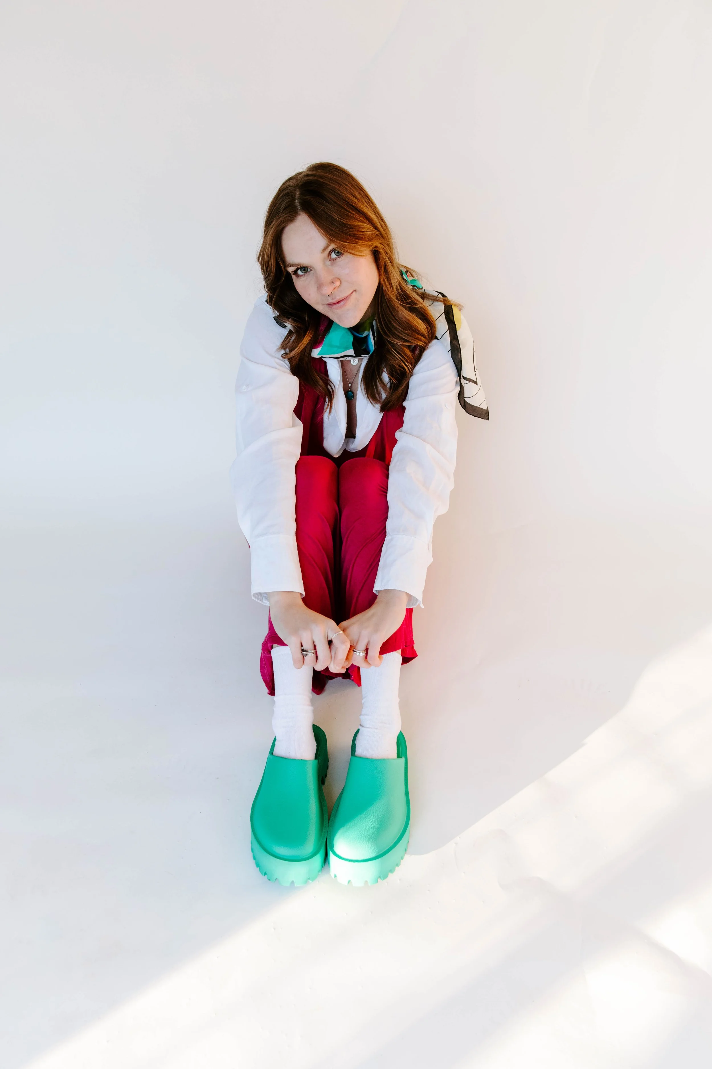 A young woman with red hair and fair skin sitting on the ground against a plain white wall, wearing a colorful scarf, pink pants, white socks, and green rain boots.