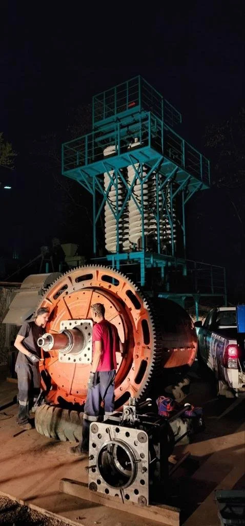 Two men inspecting a large industrial gear and mechanical machinery at night, with a tall blue metal structure in the background and tools on the ground.