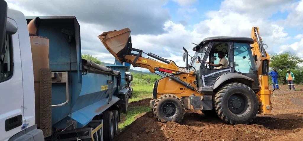 A backhoe loader is loading dirt into a truck on a construction site. Two workers are visible in the background, standing on the dirt.