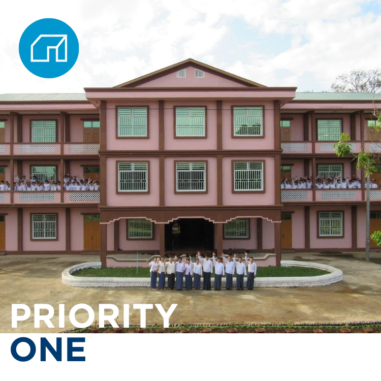 A three-story pink school building with students standing outside and on the balconies, and a group of students standing in front of the building.