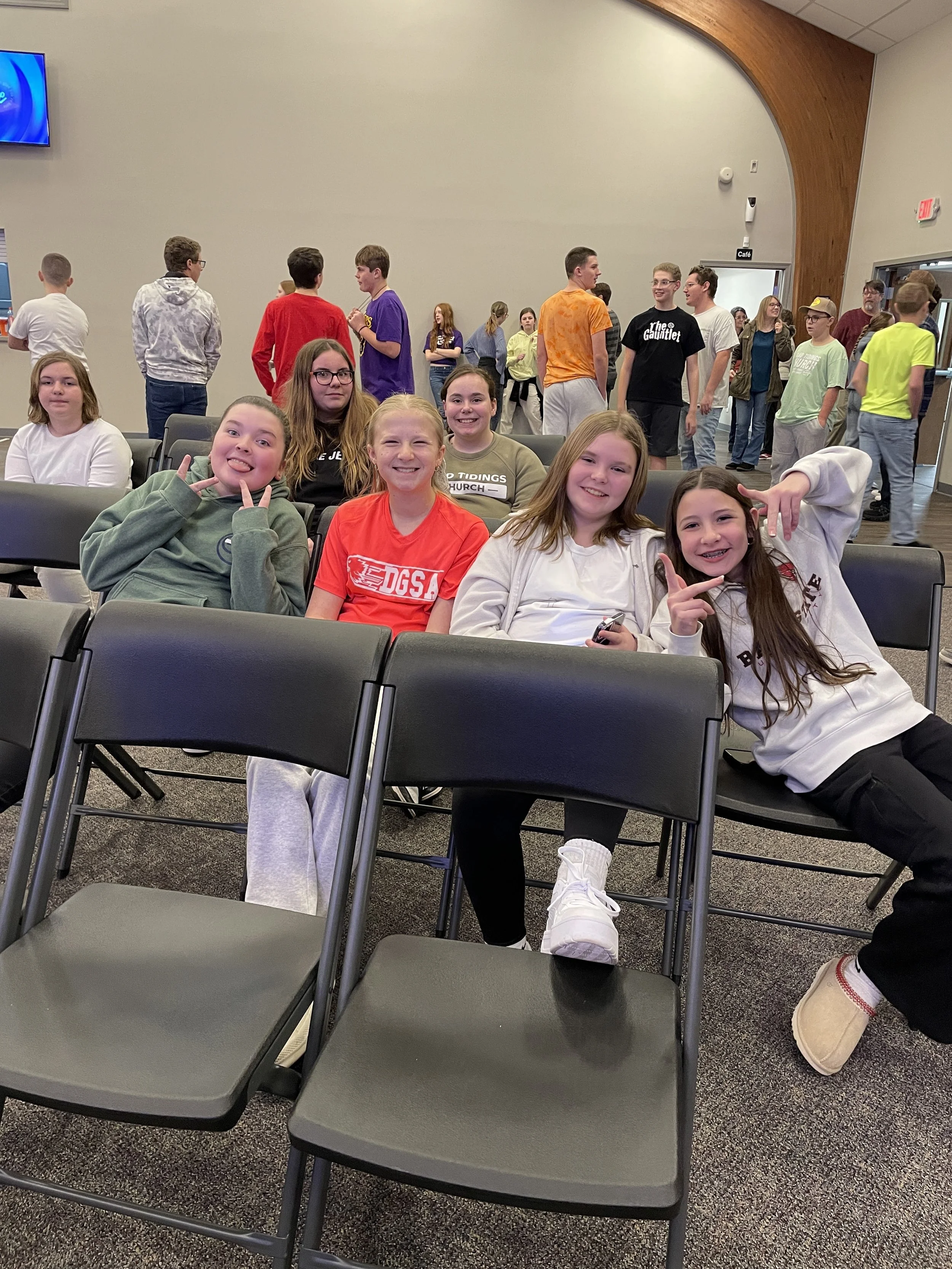 A group of girls who are friends smile and pose as other youth and students mingle in the background. 