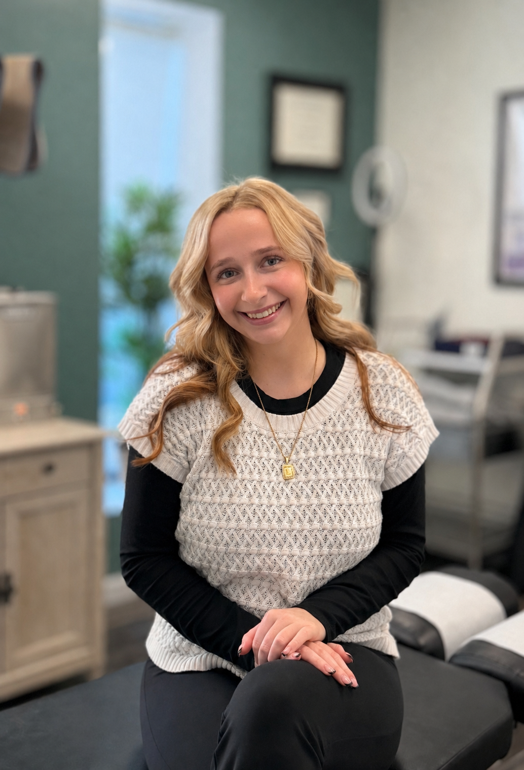 A young woman with blonde wavy hair smiling, seated in a room that appears to be a medical or professional setting, wearing a black long-sleeve top under a white knit short-sleeve sweater with a necklace.