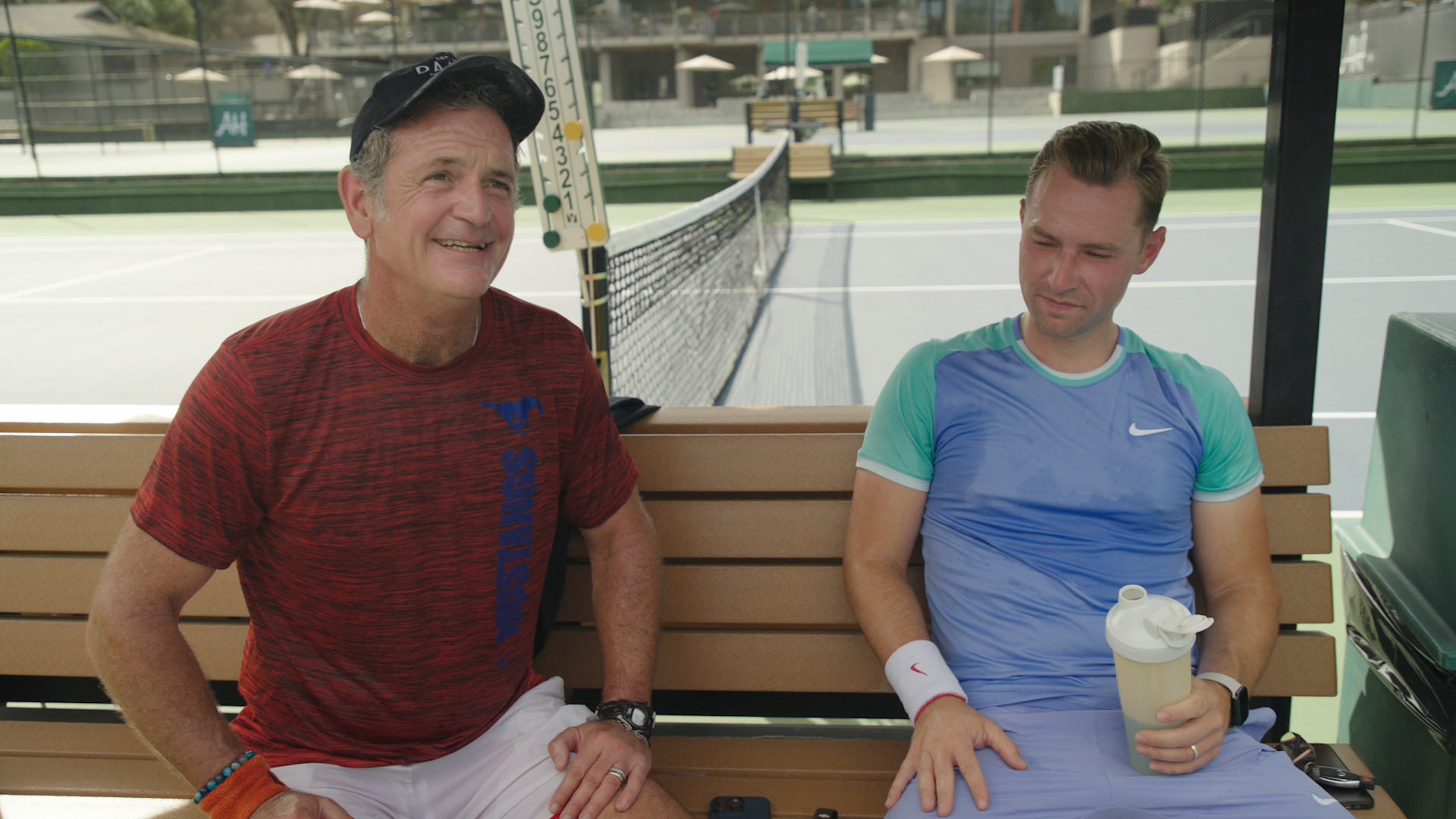 Philipp Wehn sitting beside Silicon Valley investor John Pleasants on a bench at a tennis court, both relaxing post-match, with tennis gear and court visible in the background.