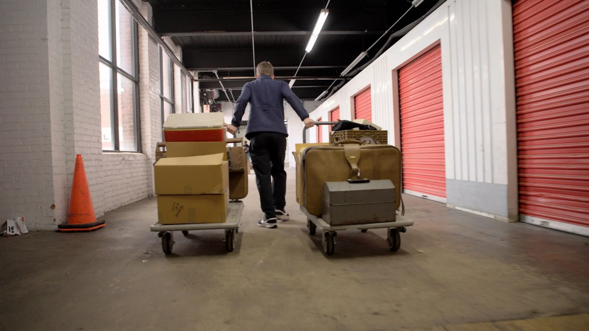 Michael pushes two loaded flat carts through a storage facility hallway lined with red doors