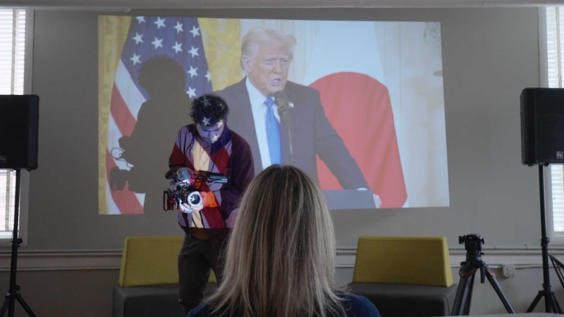 Documentary cameraman filming in front of a projected image of Donald Trump speaking at a press conference