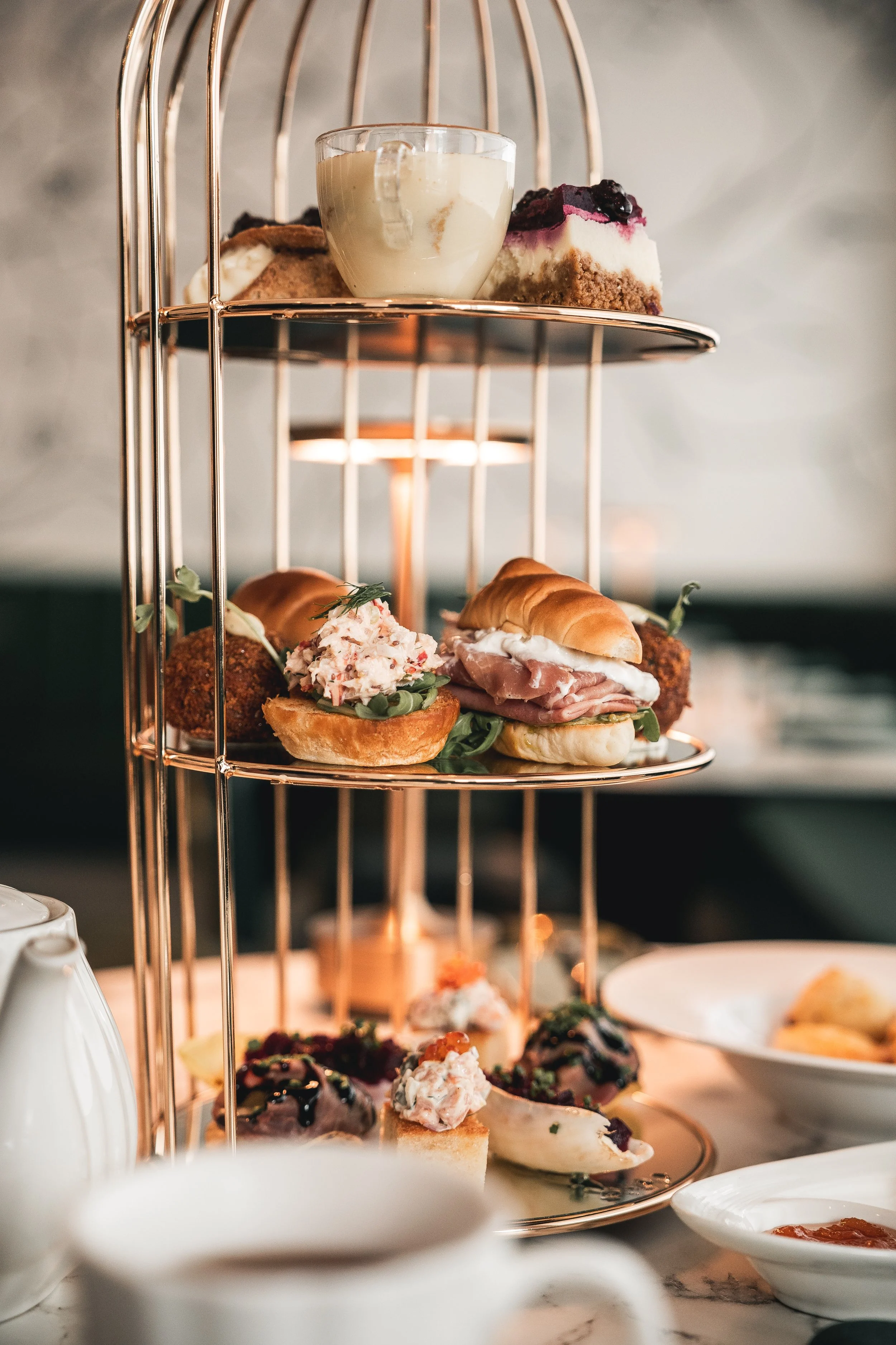 A three-tiered copper serving stand loaded with assorted sandwiches, pastries, cakes, and desserts, set on a table with teapots and plates.