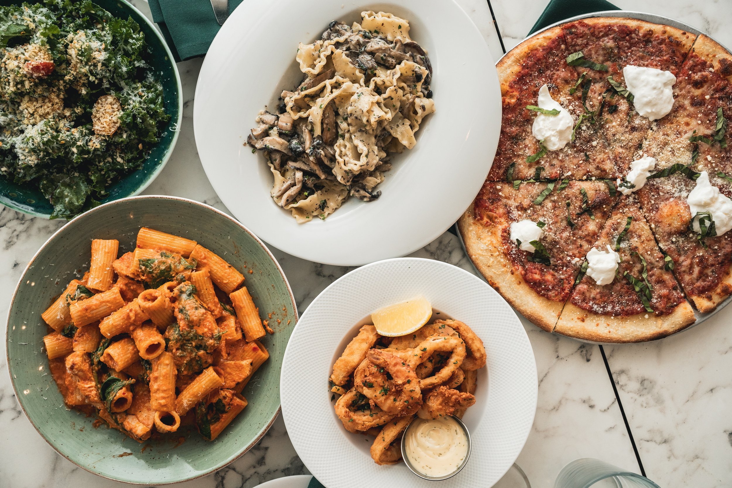An assortment of Italian dishes on a marble table, including a green bowl of Caesar salad, a white bowl of pasta with mushroom sauce, a white plate of fried calamari with lemon and dipping sauce, a green bowl of baked rigatoni with red sauce, and a large pizza with basil, mozzarella, and tomato sauce.