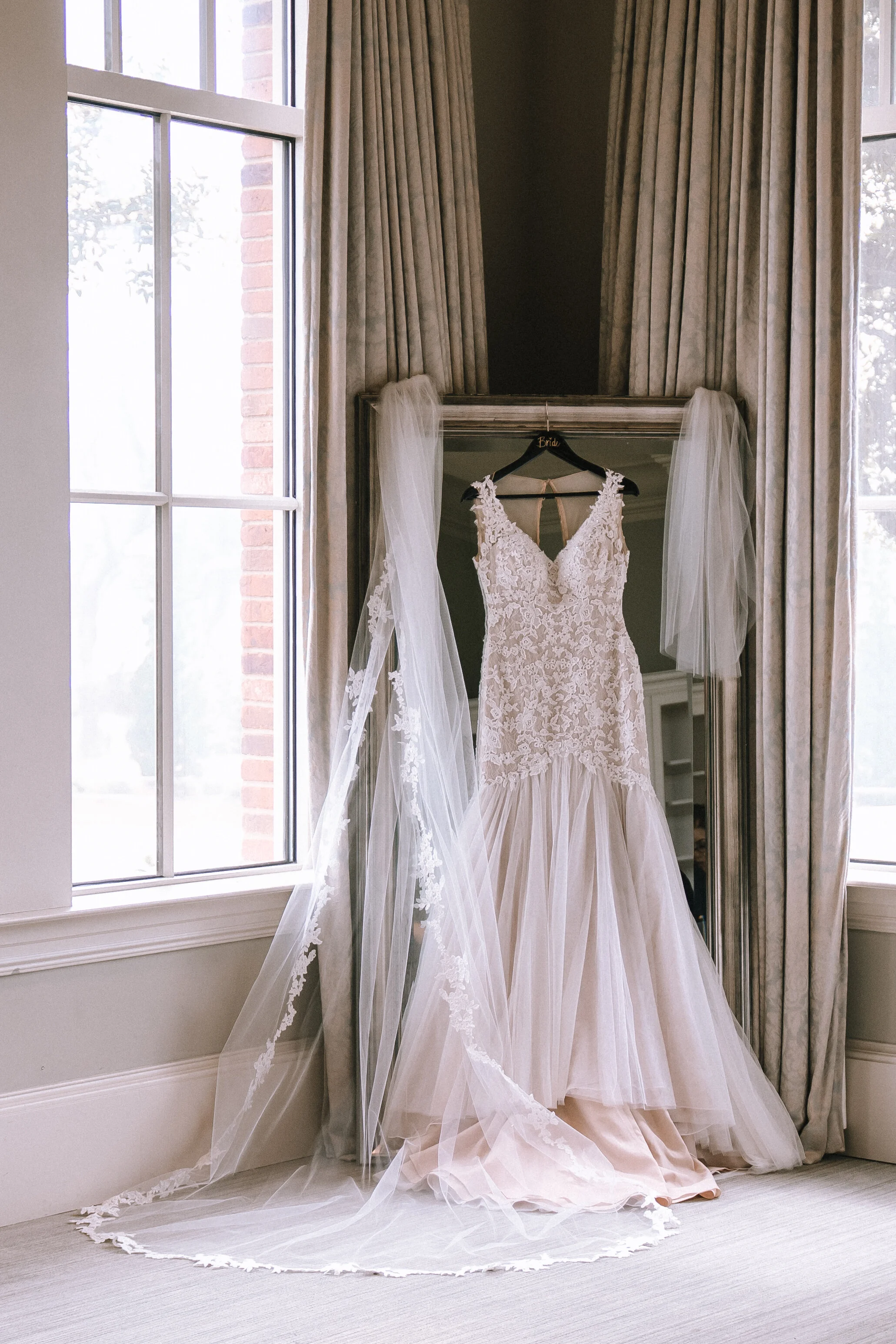 Wedding dress hanging on a mirror with a veil draped over it, in a room with large windows and curtains. Pensacola wedding photography - pensacola wedding photographer