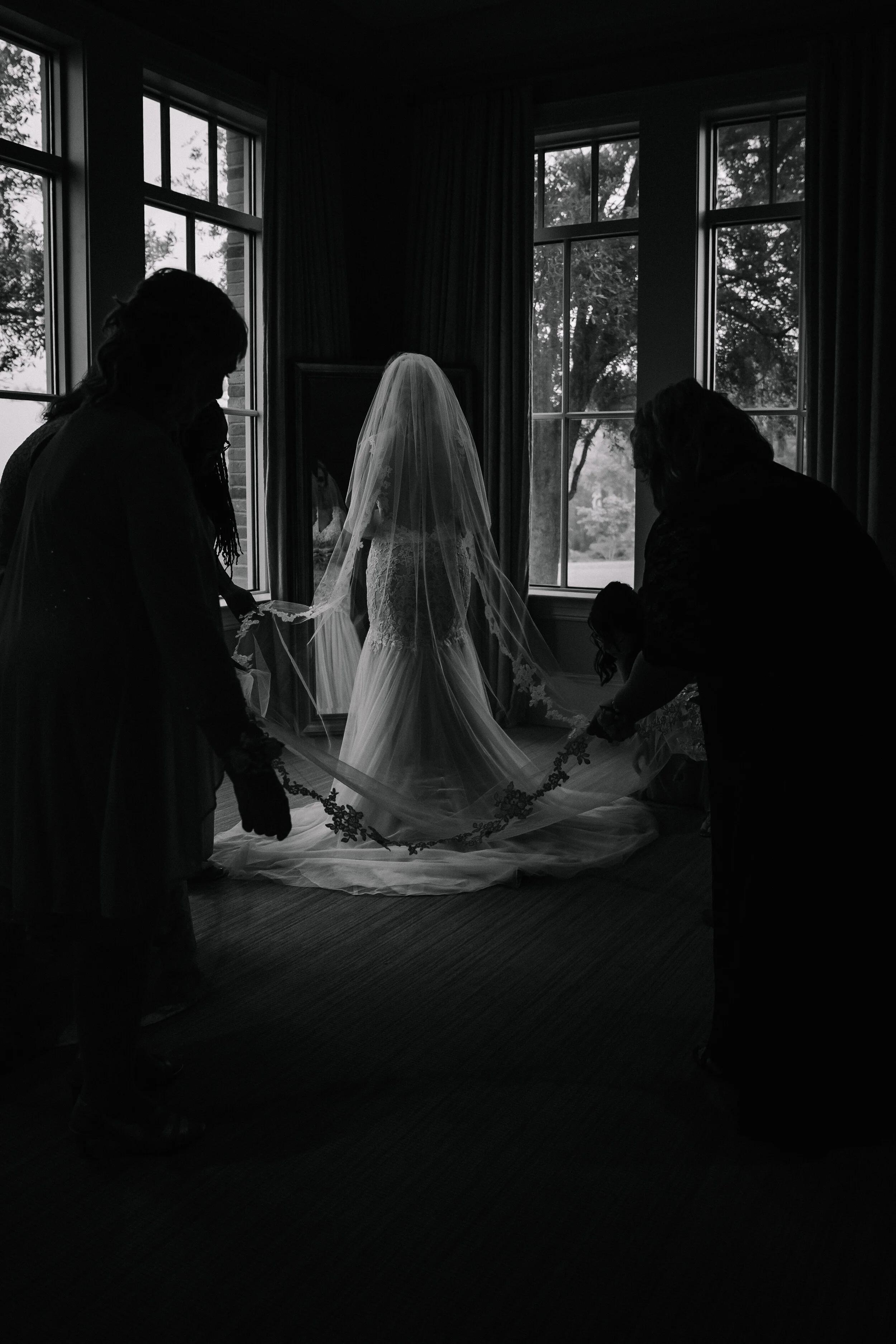 Silhouette of a bride in a wedding dress and veil standing by windows, with two women adjusting the train of her dress in a dimly lit room. pensacola wedding photography - pensacola wedding photographer