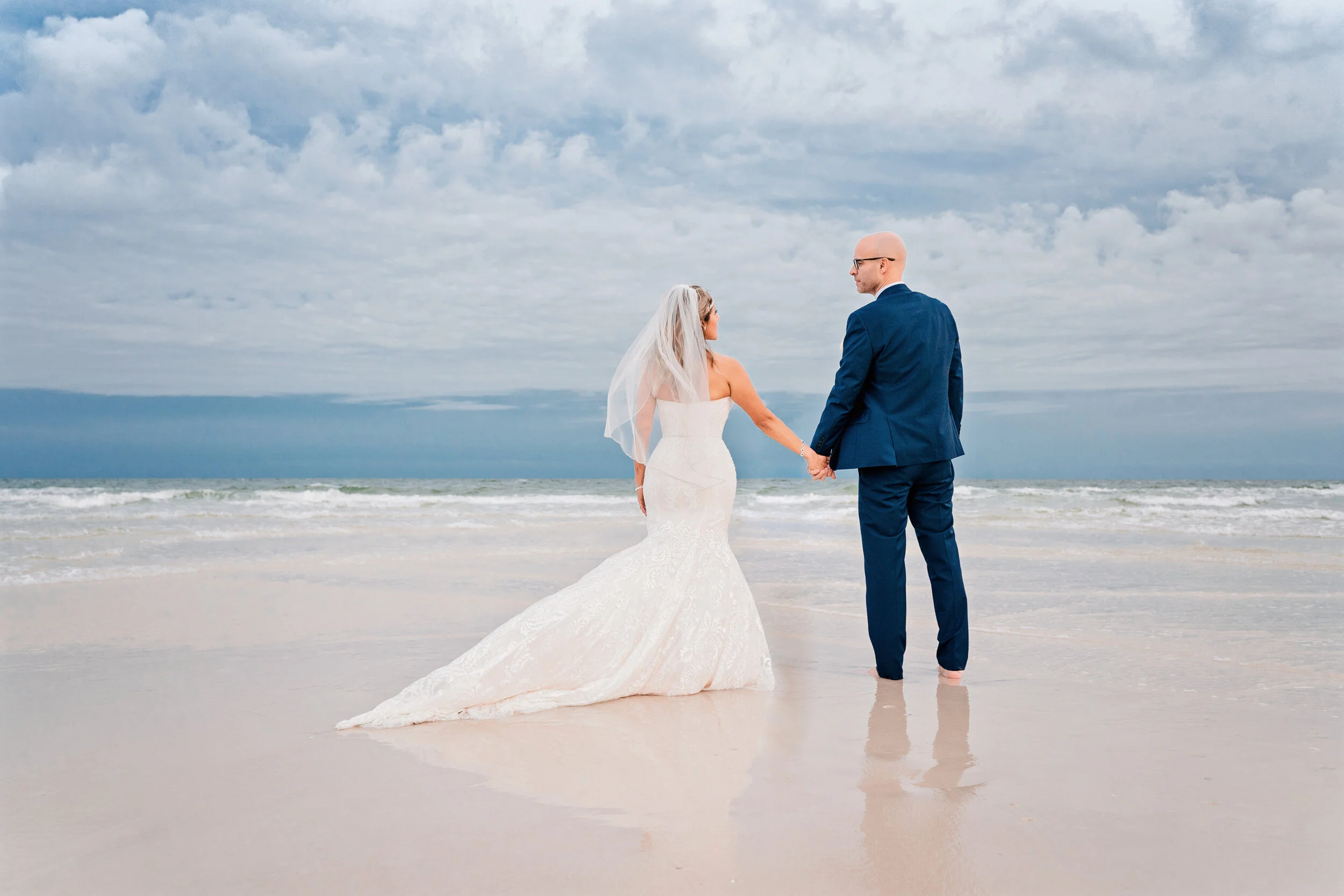 A bride and groom in wedding attire holding hands on a beach, facing the ocean with a cloudy sky overhead.Pensacola wedding photography - pensacola wedding photographer