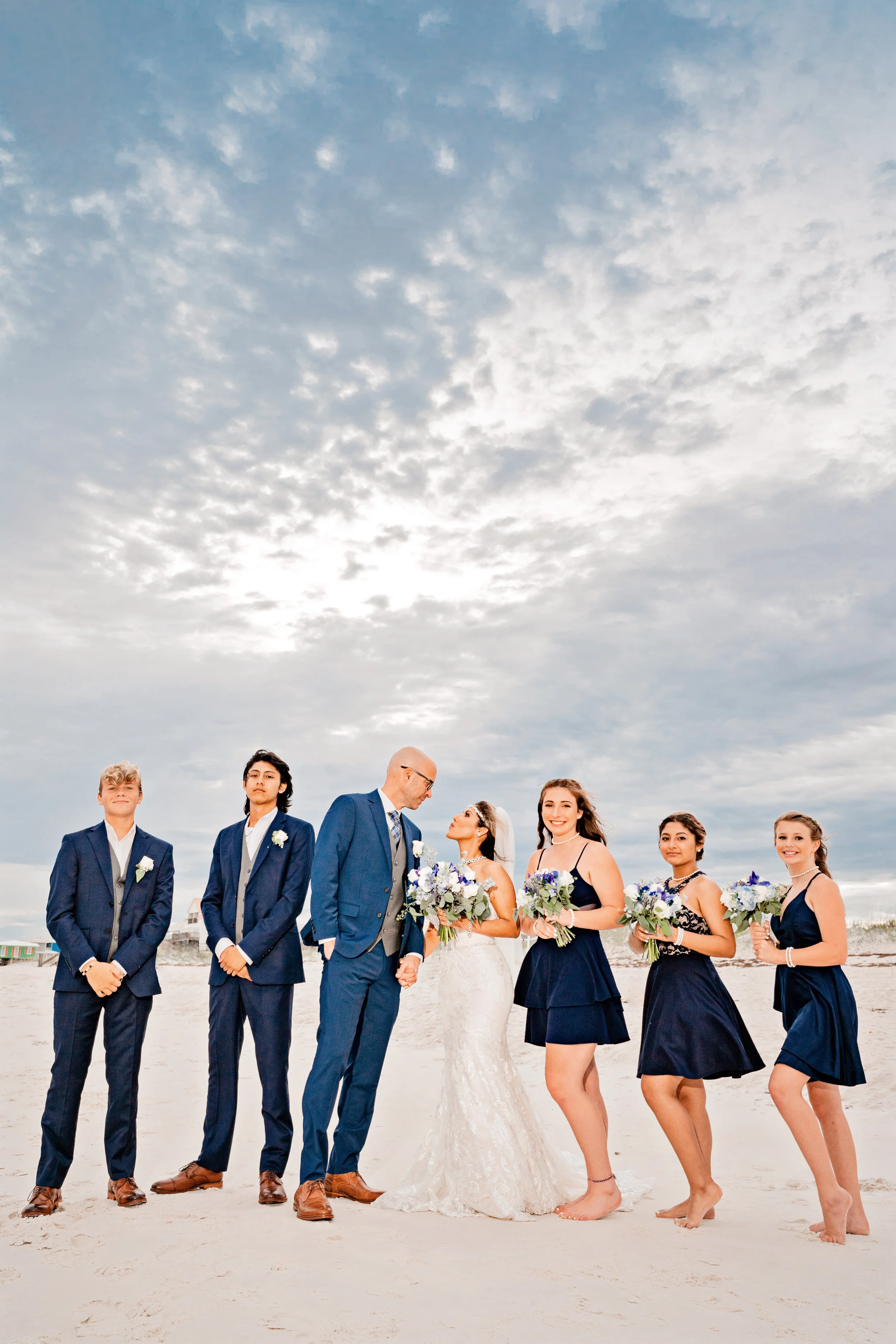 A wedding party on the beach featuring the bride in a white gown, groom in a blue suit, and bridesmaids and groomsmen in matching dark blue outfits, holding bouquets, with a cloudy sky overhead.Pensacola wedding photography - pensacola wedding photog