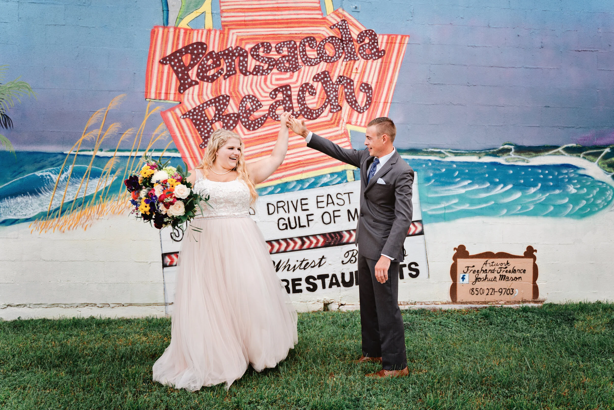 A bride and groom celebrating in front of a colorful mural that reads 'Pensacola Beach'. The bride is holding a bouquet of flowers, and they are smiling and holding hands up in celebration.Pensacola wedding photography - pensacola wedding photographe