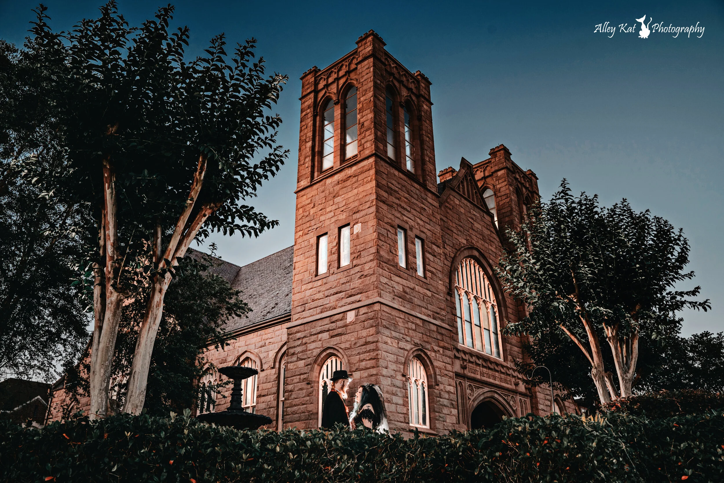 A stone church with tall gothic windows, surrounded by trees and bushes, with a couple standing in front wearing formal attire during sunset Pensacola wedding photography - pensacola wedding photographer