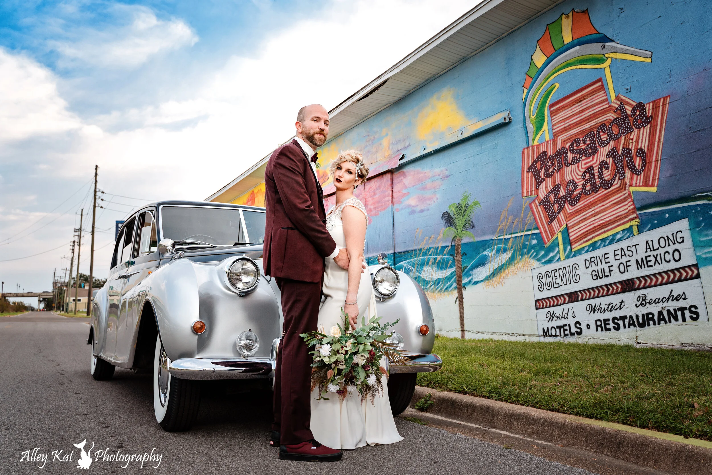 A newlywed couple standing in front of a vintage car along a mural wall reading 'Panorama Beach' with palm trees, waves, and the words 'Scenic drive east along Gulf of Mexico',Pensacola wedding photography - pensacola wedding photographer