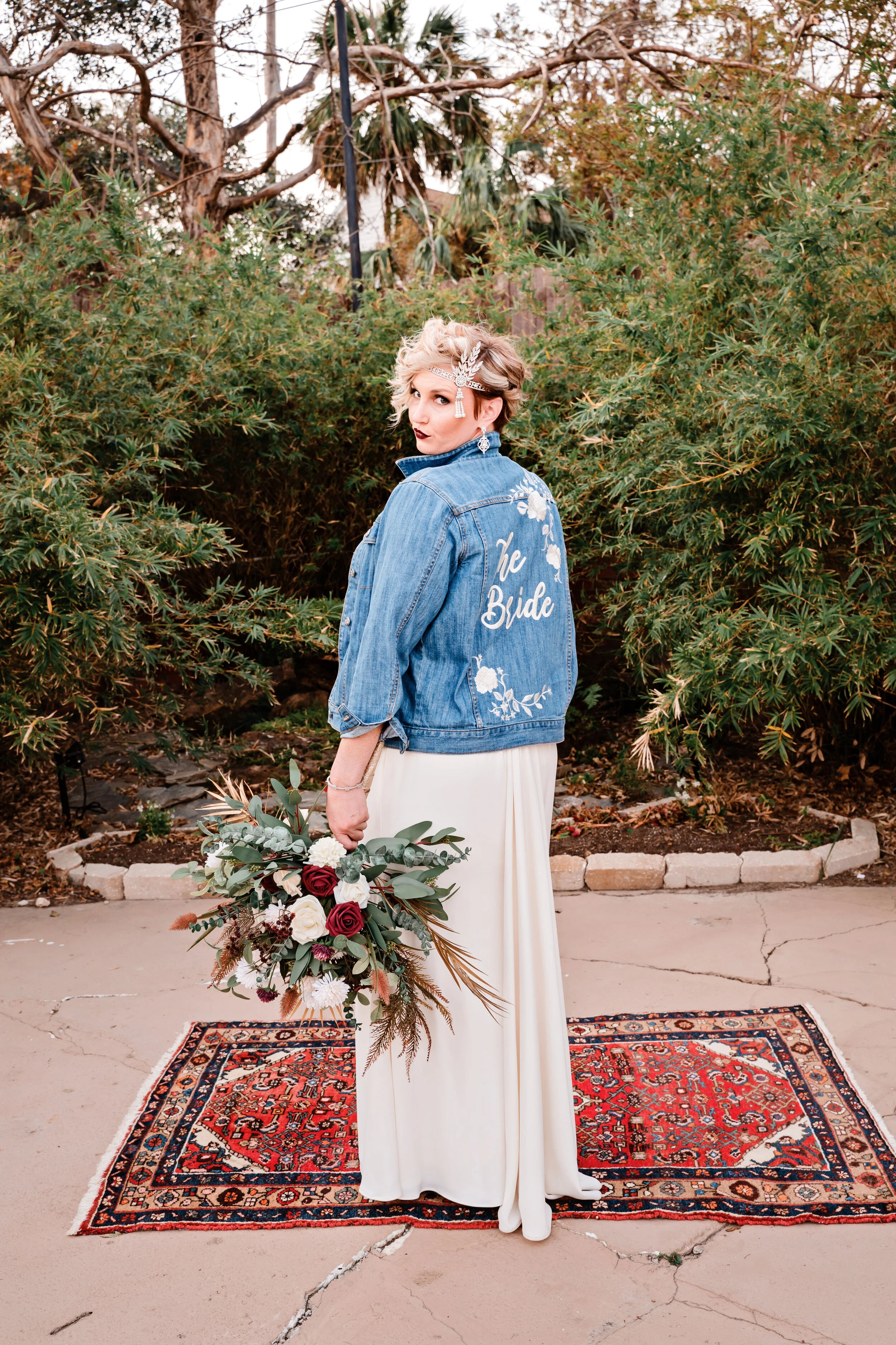 A bride with short blonde hair wearing a white dress and a blue denim jacket that says 'the Bride' on the back, standing outdoors on a decorative rug, Pensacola wedding photography - pensacola wedding photographer