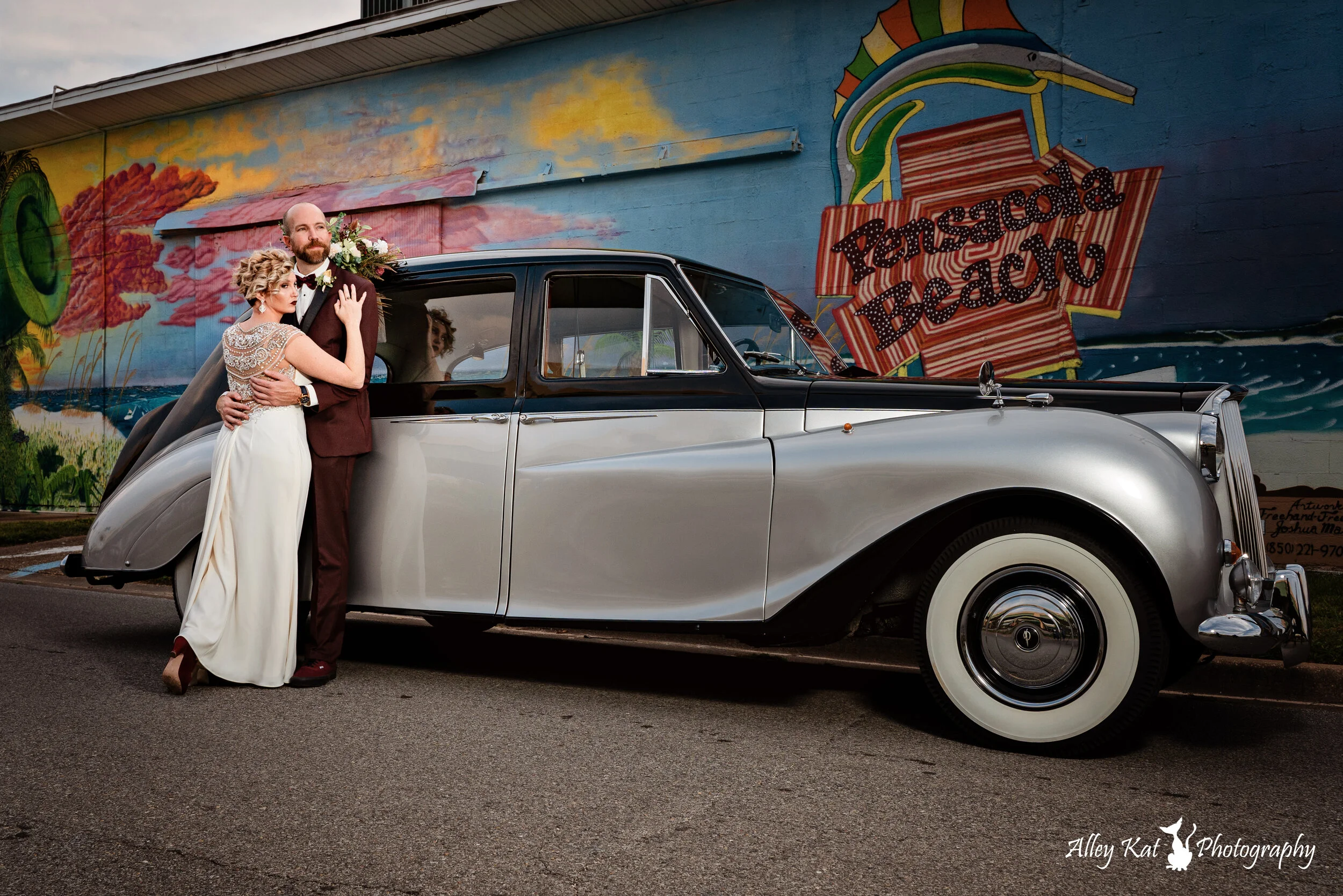 A bride and groom hugging next to a vintage silver and black car, with a colorful mural of a beach scene and the words 'Pensacola Beach' painted on the wall behind them.