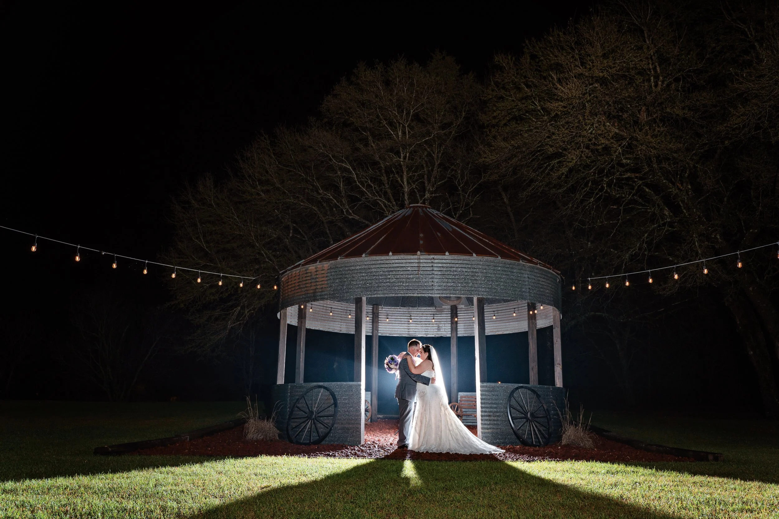 A bride and groom embrace and kiss at night under a pavilion with string lights, illuminated by a bright light behind them, on a grassy area with trees in the background.