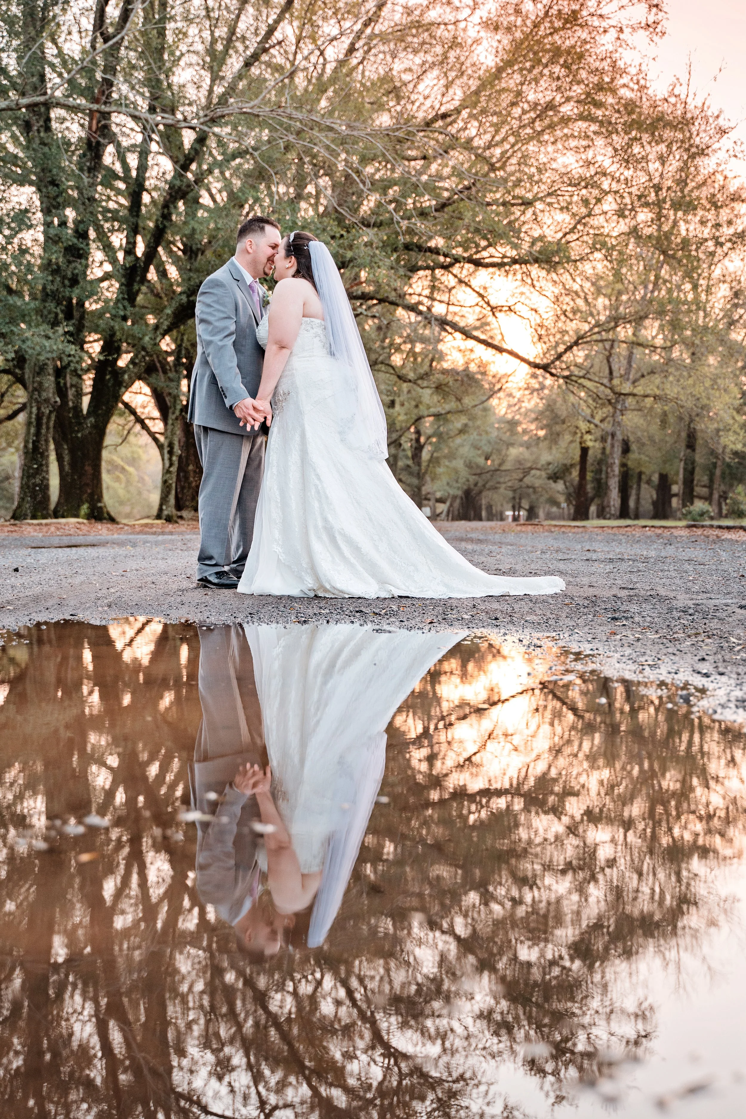 A bride and groom holding hands and touching foreheads, standing outdoors at sunset with trees in the background, with their reflection visible in a large puddle of water. Pensacola wedding photography - pensacola wedding photographer