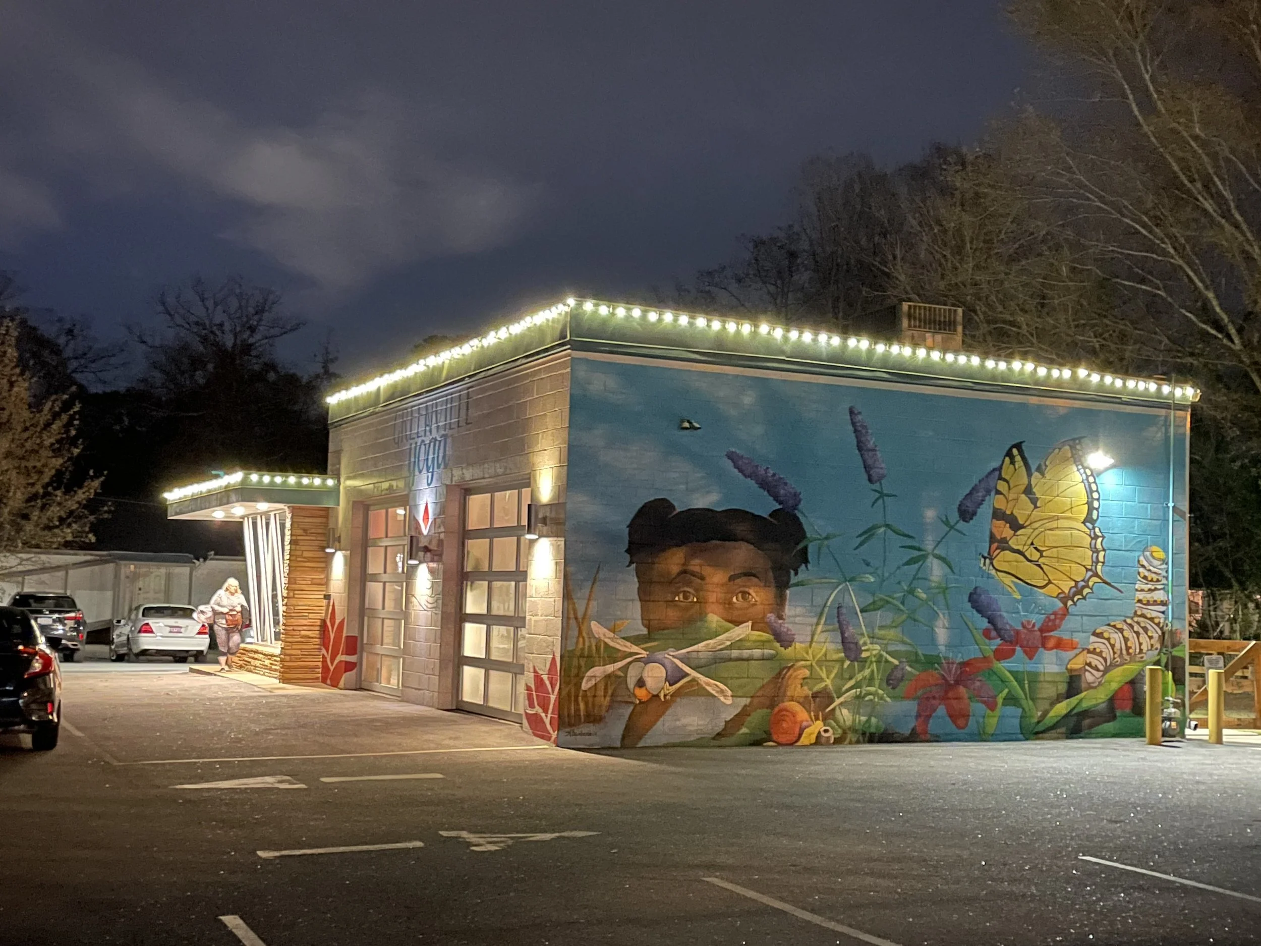 A building with colorful mural art of a child's face, butterflies, flowers, and greenery, illuminated by string lights along the roofline, with a parking lot and trees in the background during night time.