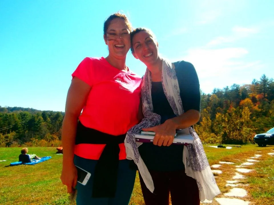 Two women smiling outdoors in a sunny field with trees in the background, one holding a folder and the other holding a smartphone.
