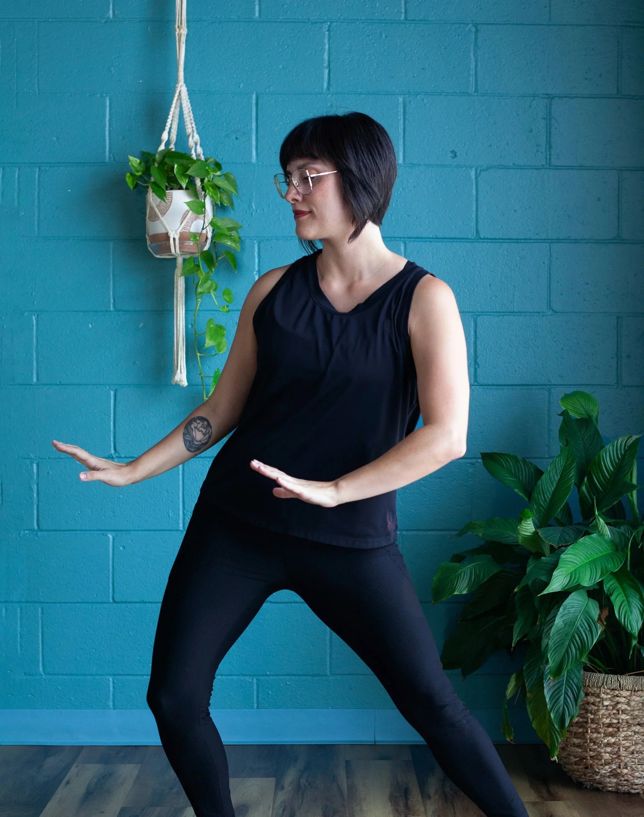 A woman with short dark hair and glasses practicing yoga indoors against a teal brick wall, with a hanging plant and a large potted green plant nearby.