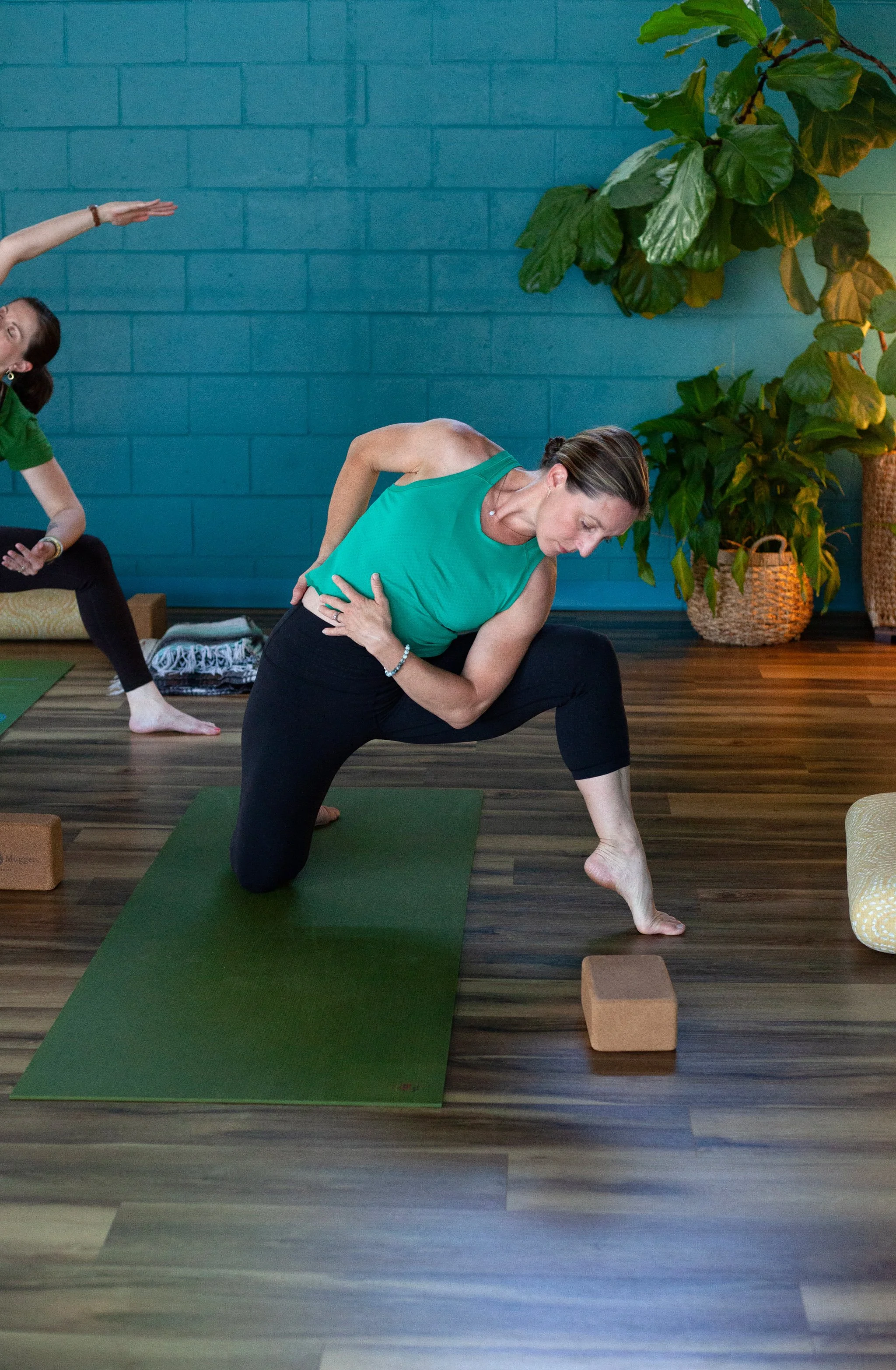 Woman in green tank top performing a yoga pose on a green mat in a wellness studio with wooden floors and a blue brick wall, near large green plants.