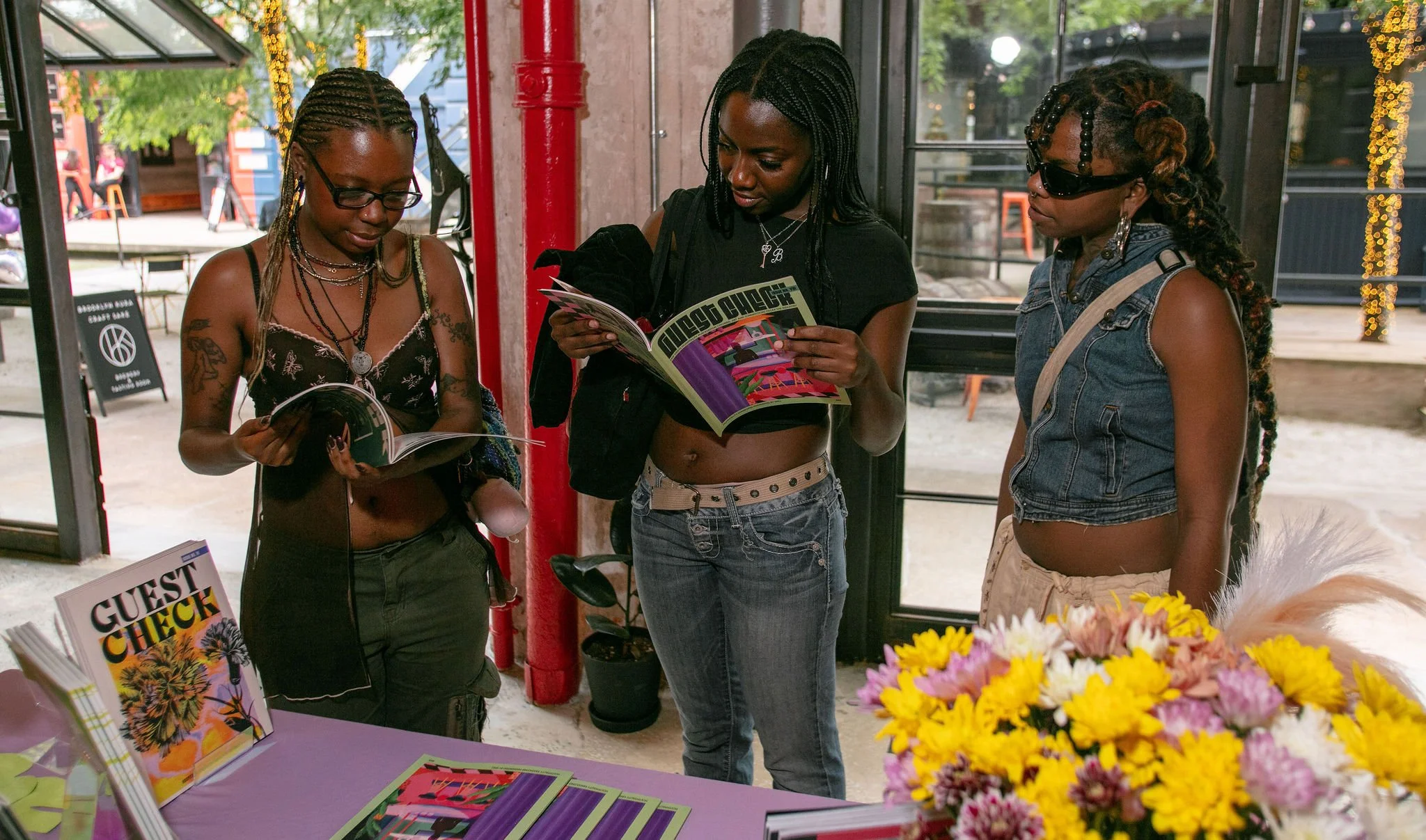Three women standing together inside a bookstore or cafe, looking at magazines, with books on a purple table and a bouquet of yellow and pink flowers in the foreground.