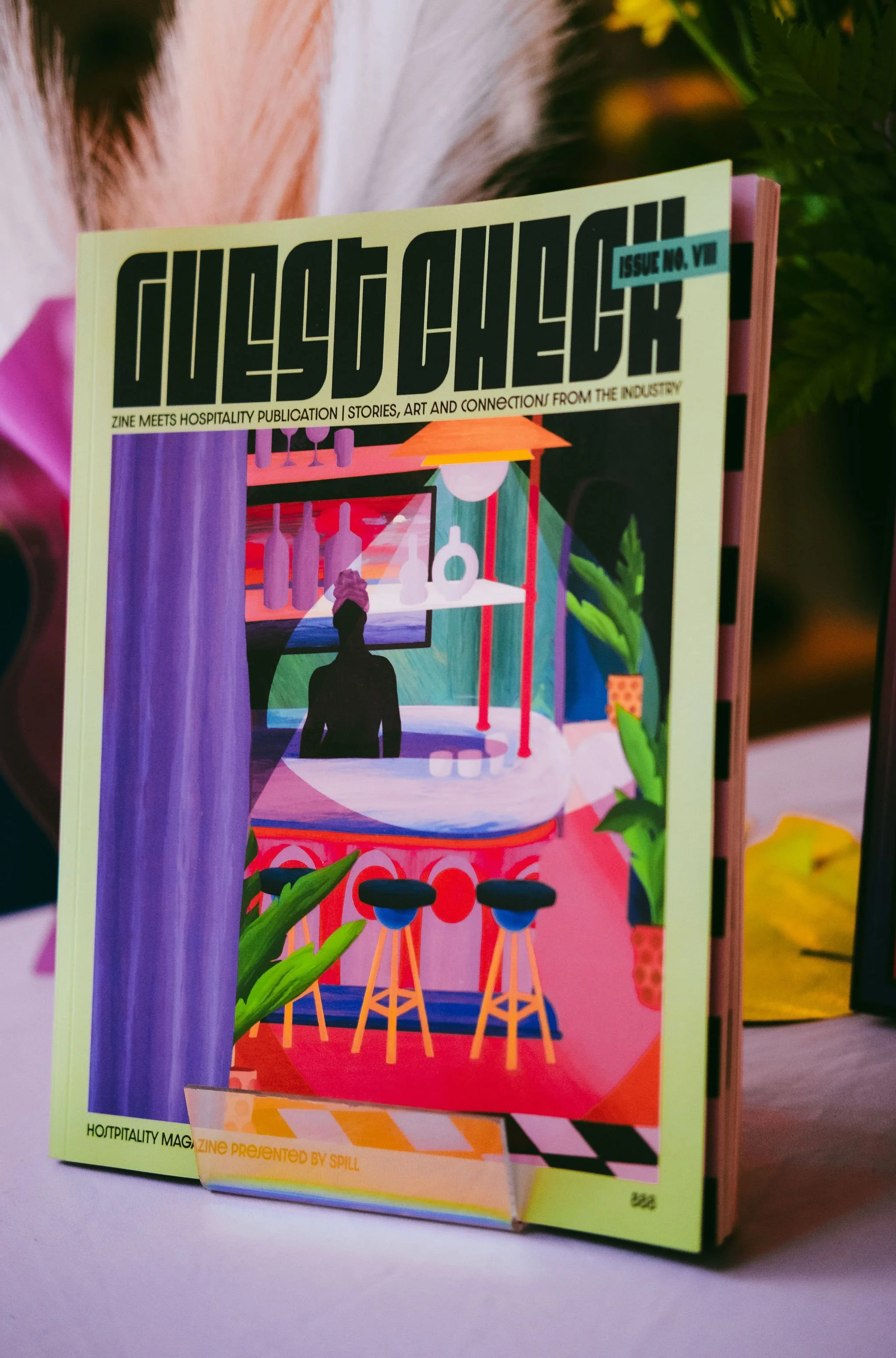 Colorful illustrated magazine cover featuring bar and lounge scene with bar stools, bottles, woman with towel turban, plants, and abstract background.