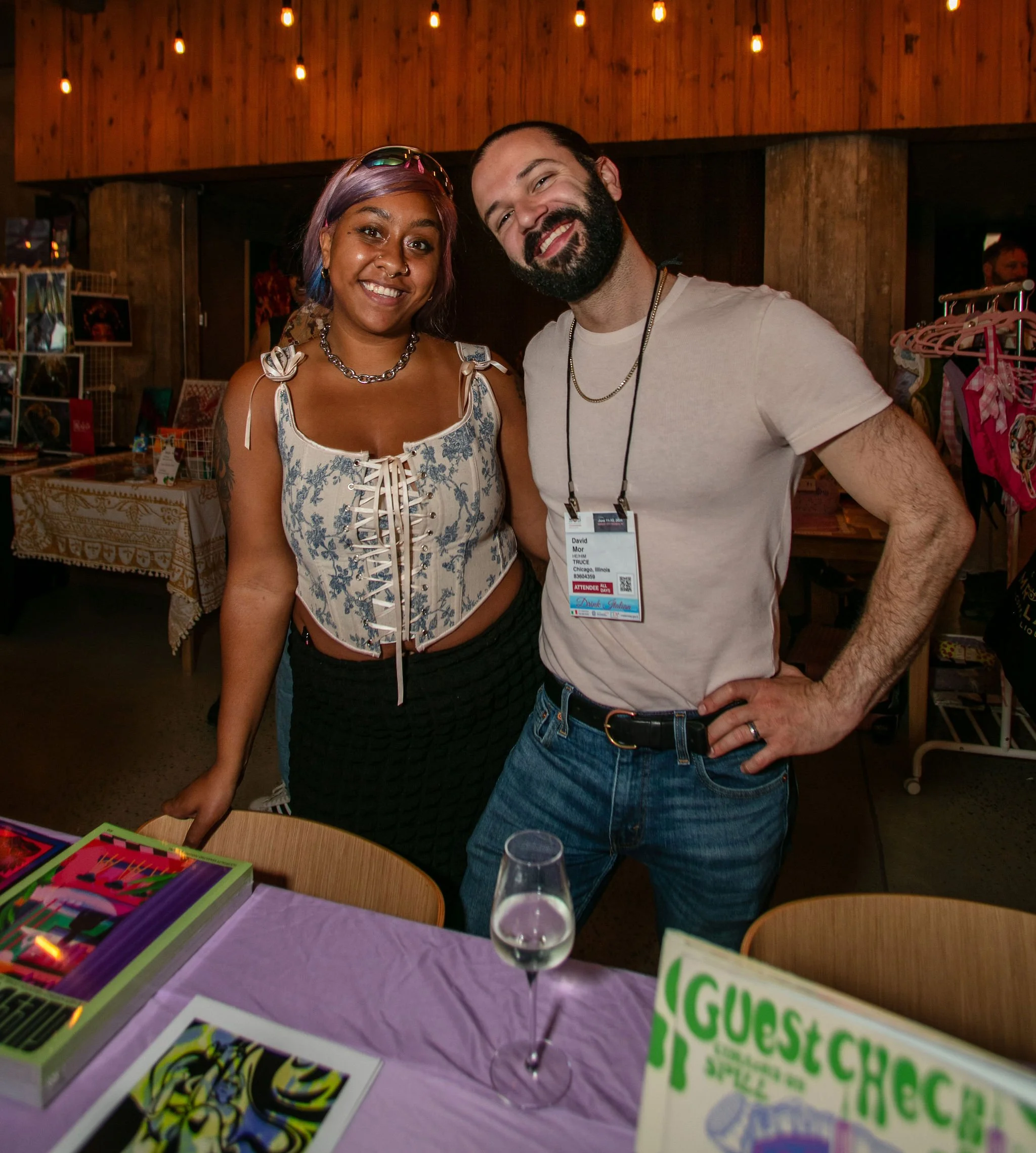 Two smiling people standing behind a table at an indoor event, with colorful items and a sign that reads "Guest Check" on the table.