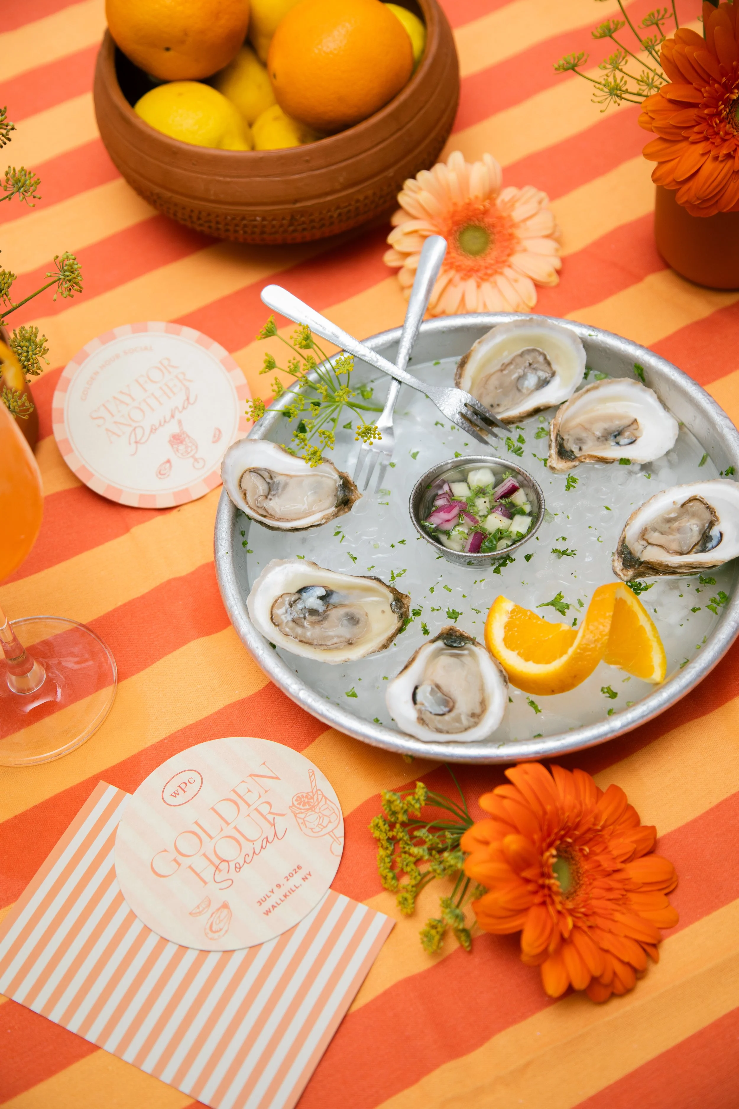 Custom Golden Hour Social coasters and napkins in peach and coral, styled on a striped tablecloth with oysters and citrus. Event print design by Studio Pierson.