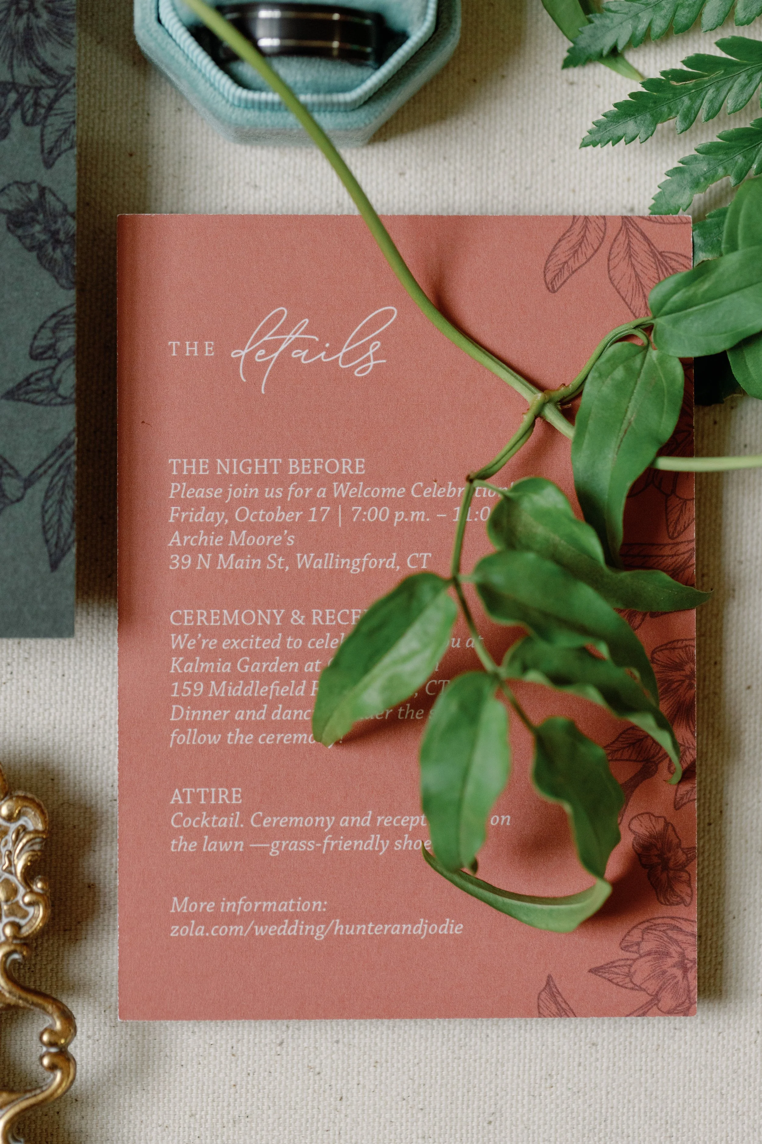 Terracotta wedding details card with white lettering and botanical illustration, styled with a wedding band and greenery. Custom wedding stationery in Connecticut.