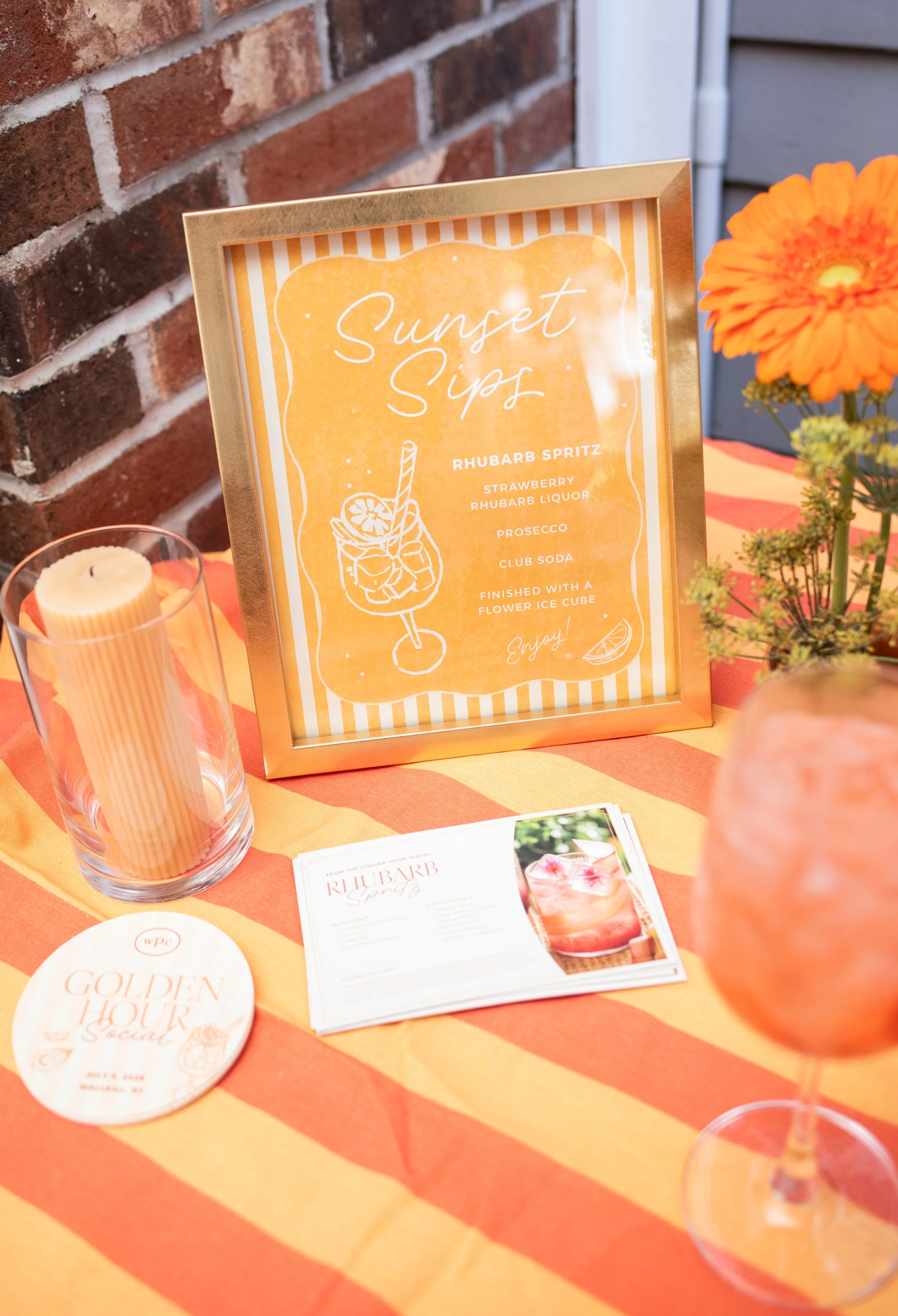 Framed Sunset Sips cocktail sign in peach and coral with a hand-drawn spritz glass illustration, listing the rhubarb spritz recipe at the Golden Hour Social. Event signage by Studio Pierson.