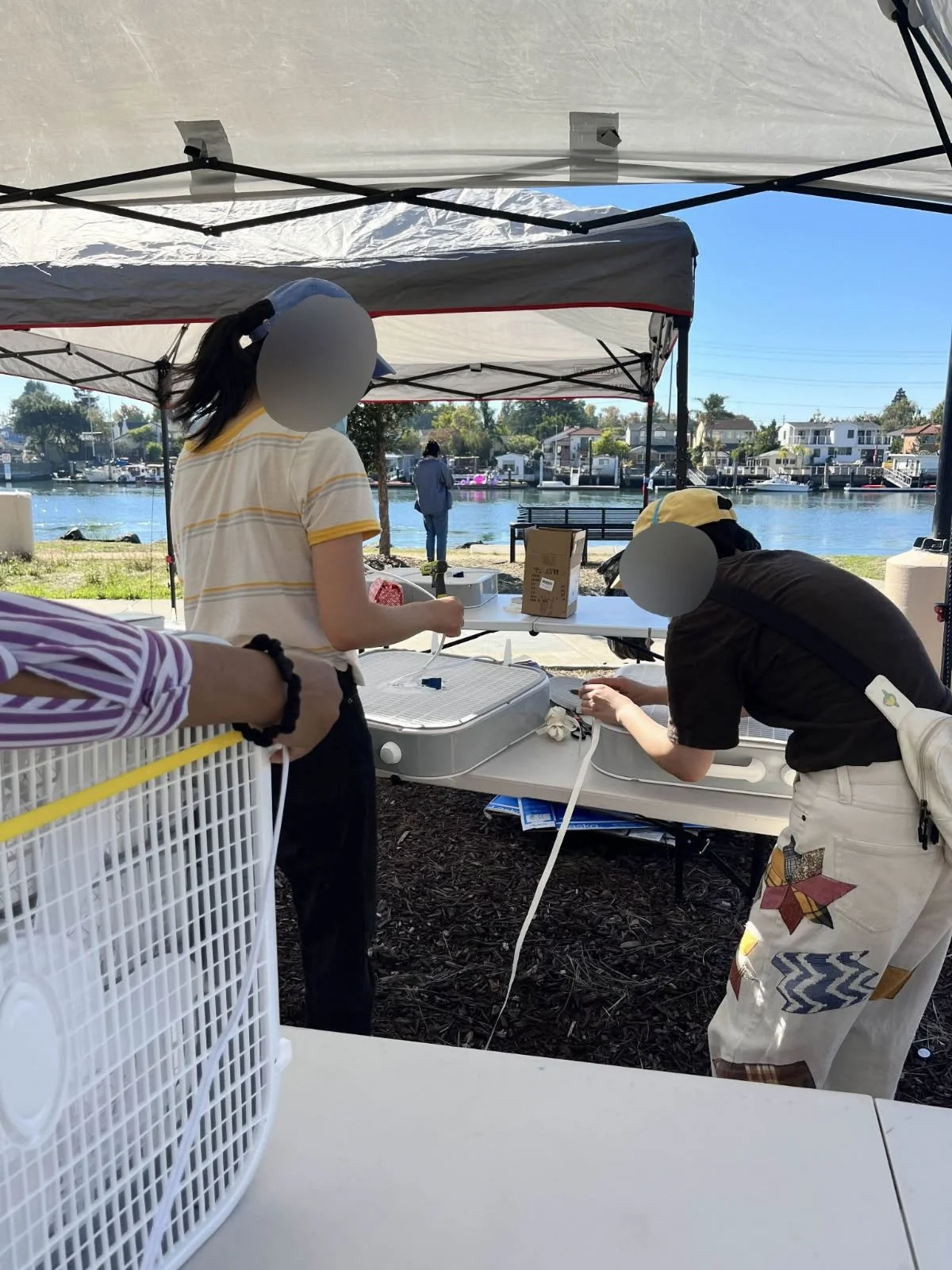 There are two people wearing masks and baseball hats in front of a table. They each have a box fan in front of them, and they are actively applying weatherstripping to the perimeter of the fans. In the background is a canopy with a second table under