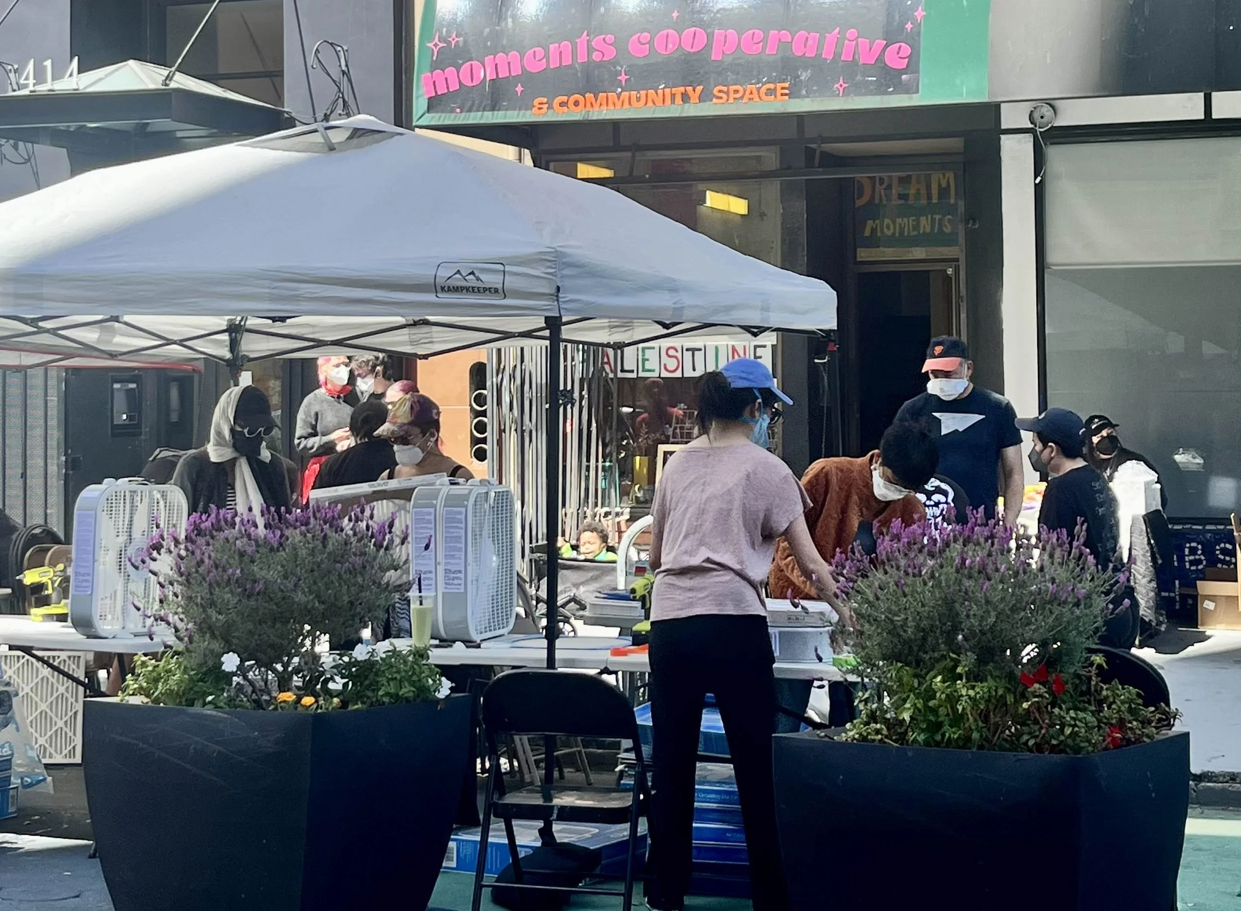 Masked people building DIY air purifiers, partially underneath a canopy, partially to the side, all in front of Moments Cooperative & Community Space.