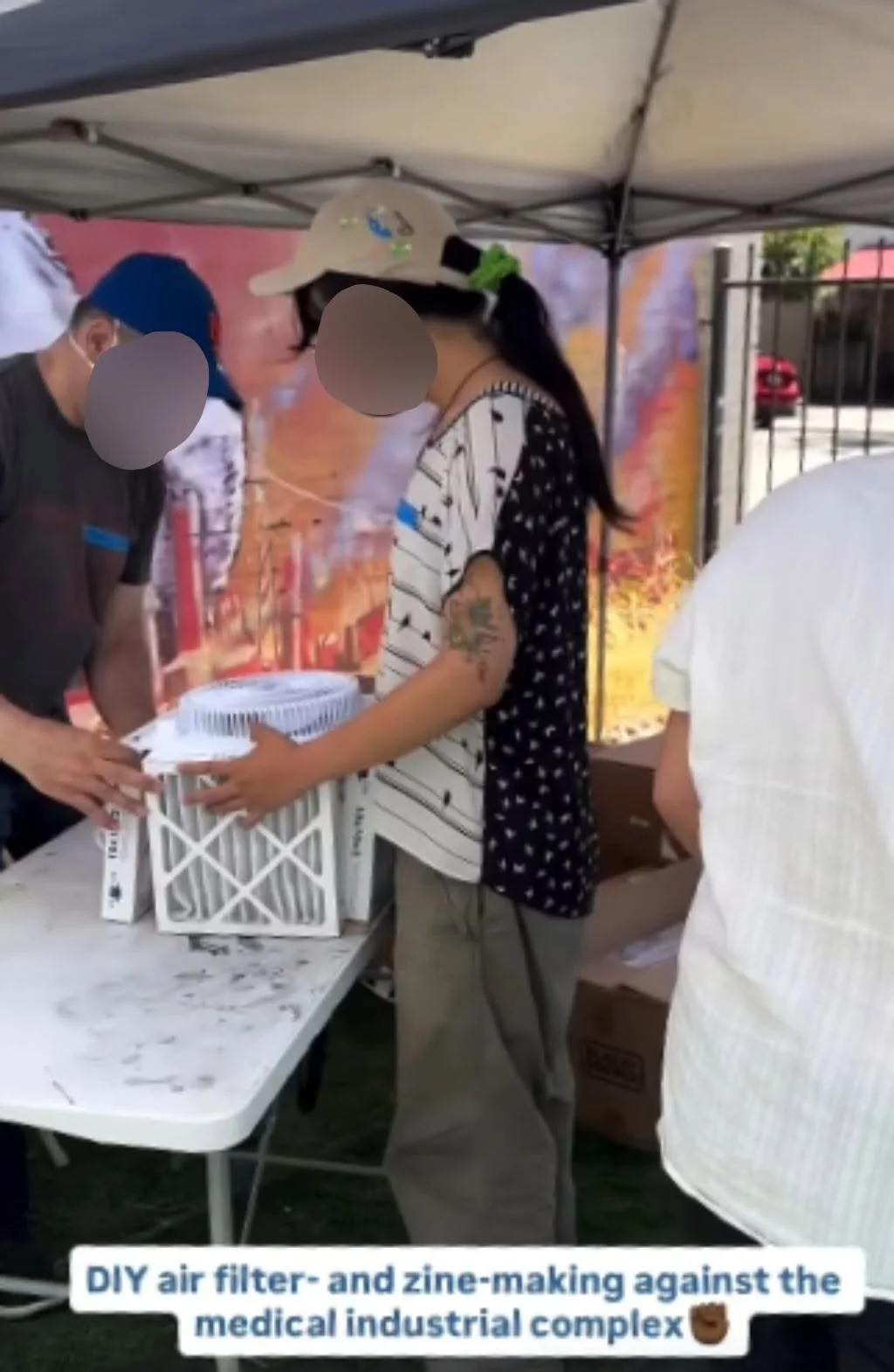 Two people wearing masks and baseball hats work together to assemble a mini CR box on a table (from LavNix's Drop the MIC event). There's a person out of frame behind them and a canopy above them. There's text at the bottom of the image that reads "D