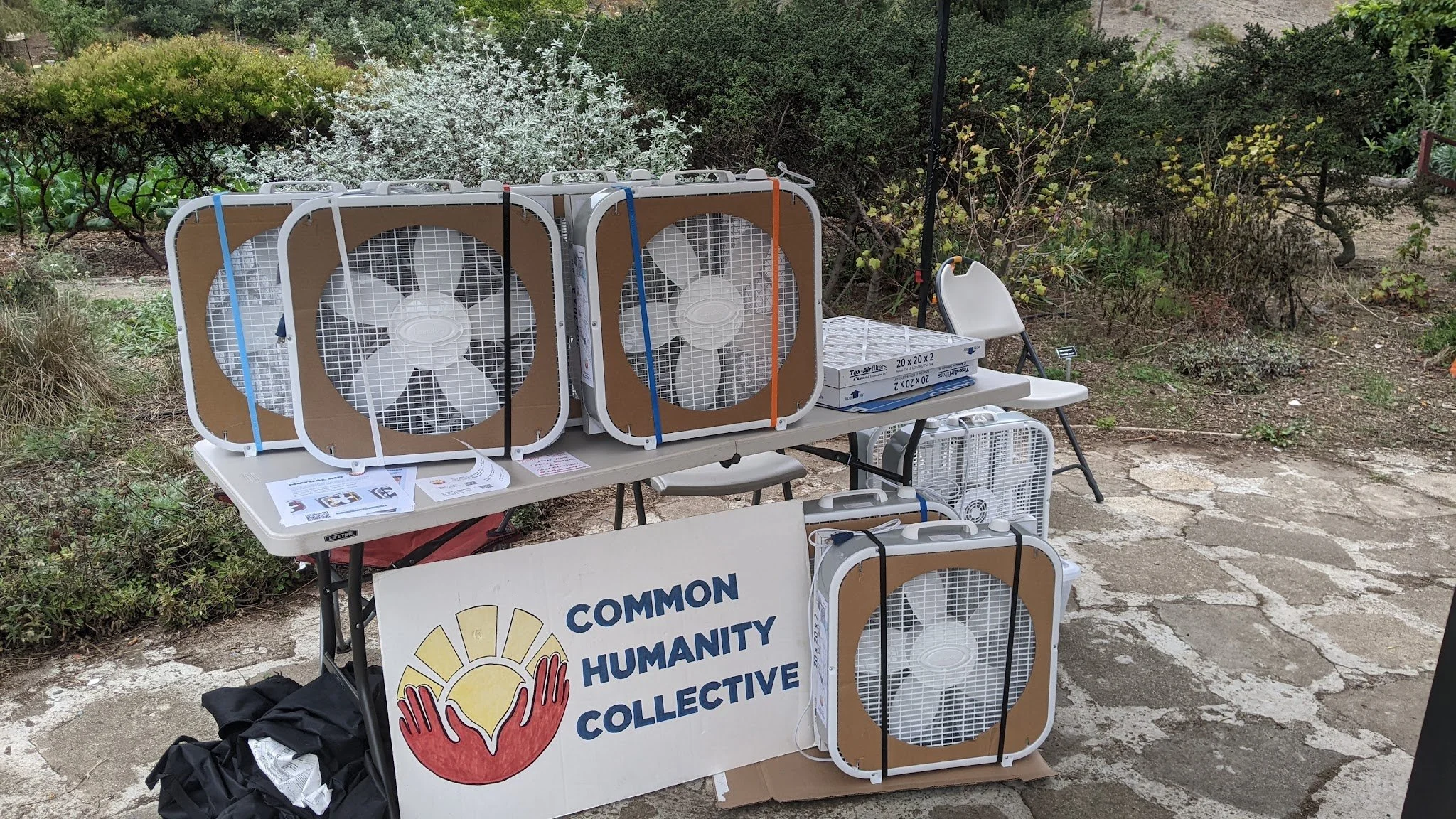 There is a table with three assembled DIY air purifiers sitting on top to the center and left, with supplies to assemble another on the right. Beneath the table is a cardboard sign that reads "Common Humanity Collective" with another assembled DIY ai